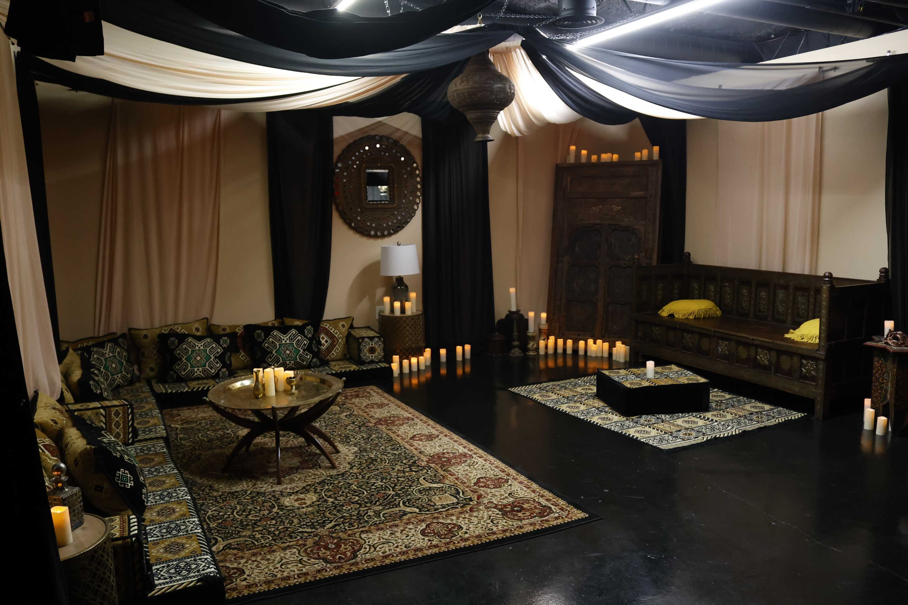 The scene features a dimly lit lounge area with patterned seating, a low table, candles on the floor, and decorative textiles hanging from the ceiling.