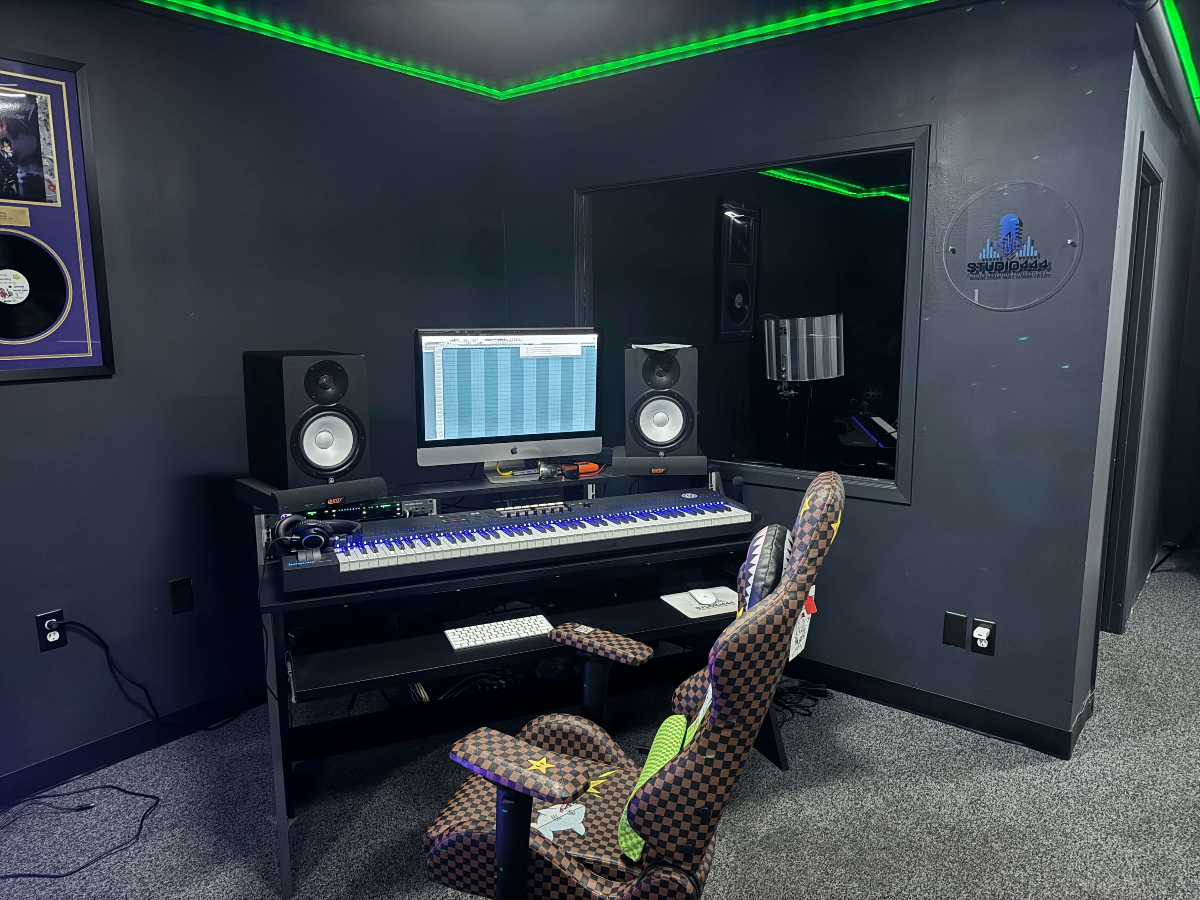 The image shows a music production studio featuring a keyboard, a computer monitor with a digital audio workstation displayed, and speakers on either side, with a decorated chair in front of the desk.