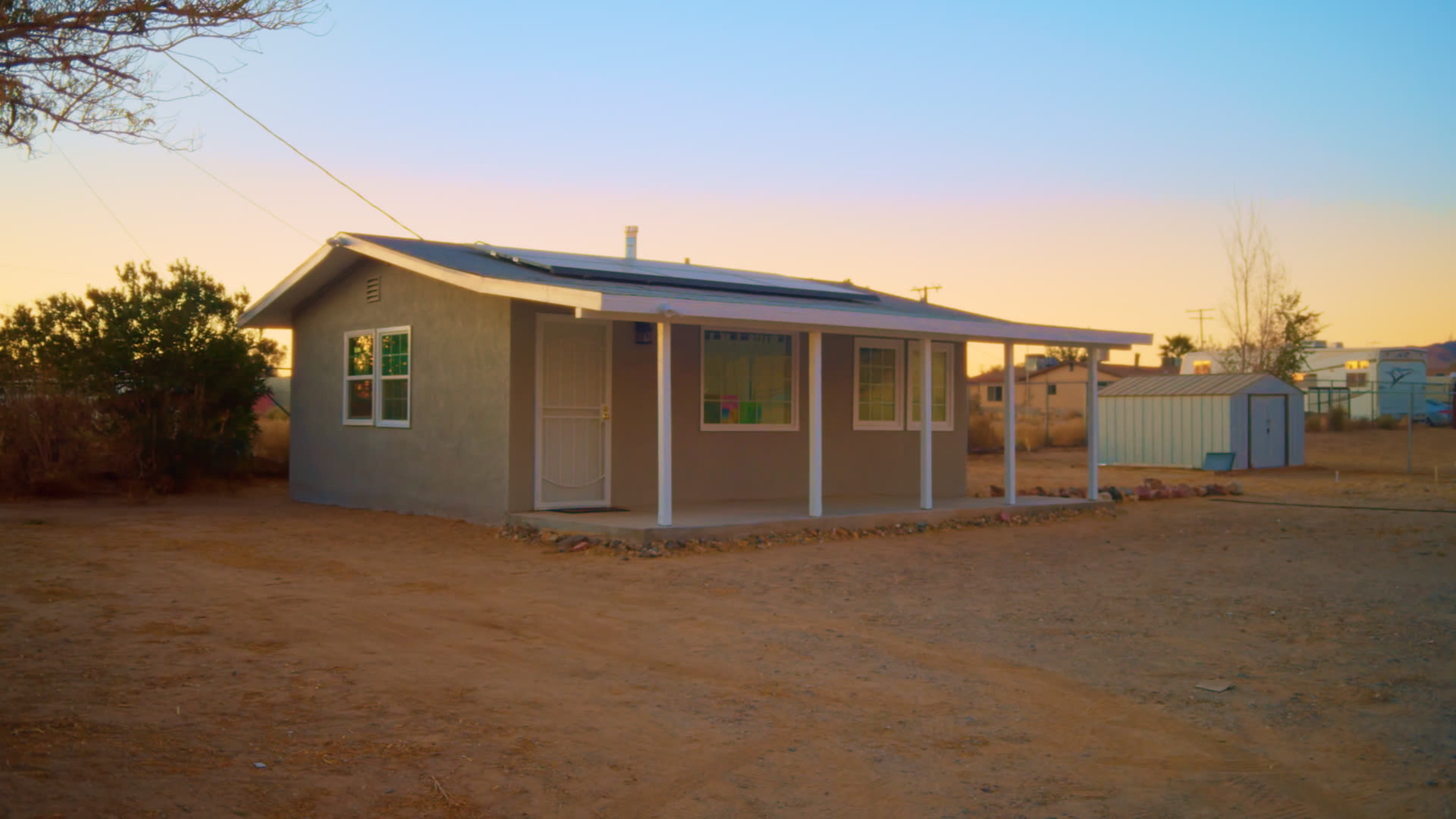 A single-story gray house with a porch and solar panels is situated on a dirt plot surrounded by sparse vegetation at dusk.