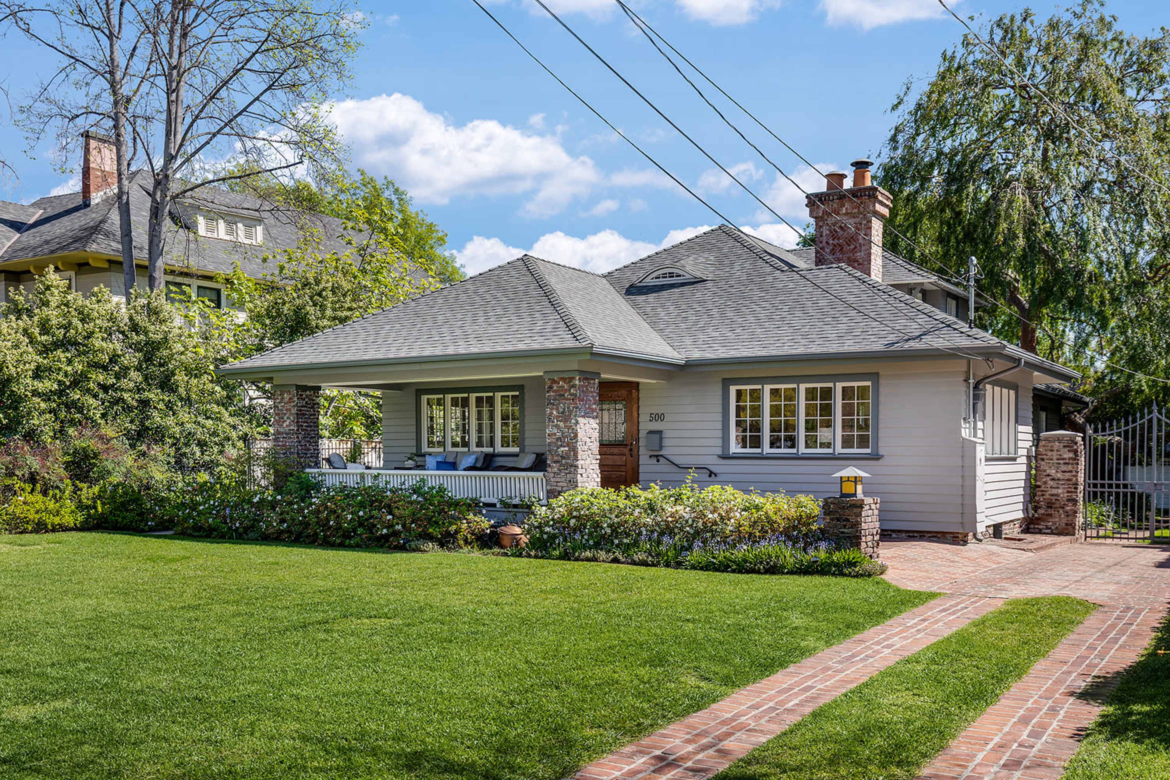 The image depicts a well-maintained single-story house with a covered front porch, surrounded by a lawn and garden, and features a brick walkway leading to the entrance.