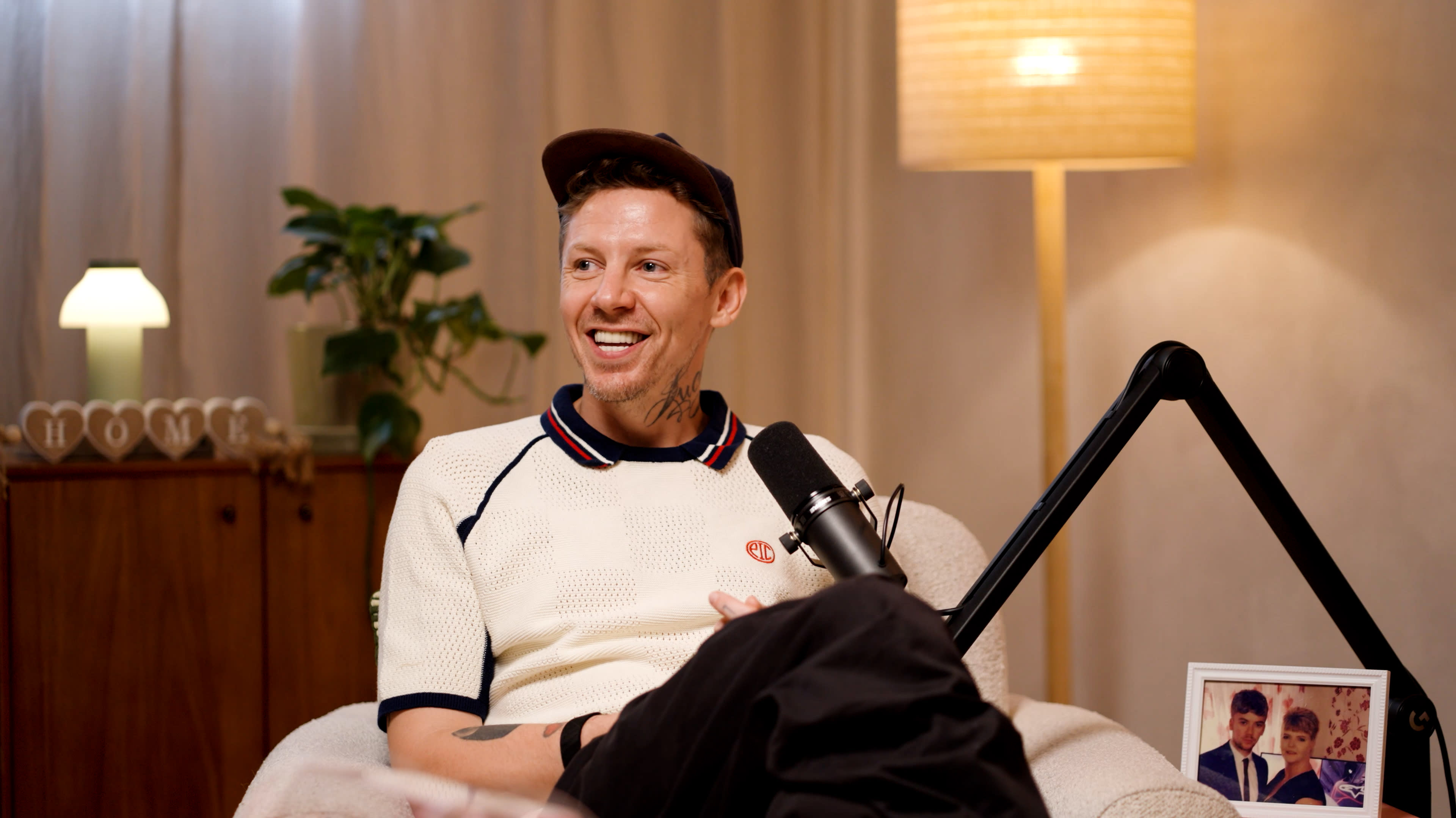 A man sits on a couch in a well-lit room, smiling and engaged in conversation, with a microphone positioned in front of him and a plant nearby.