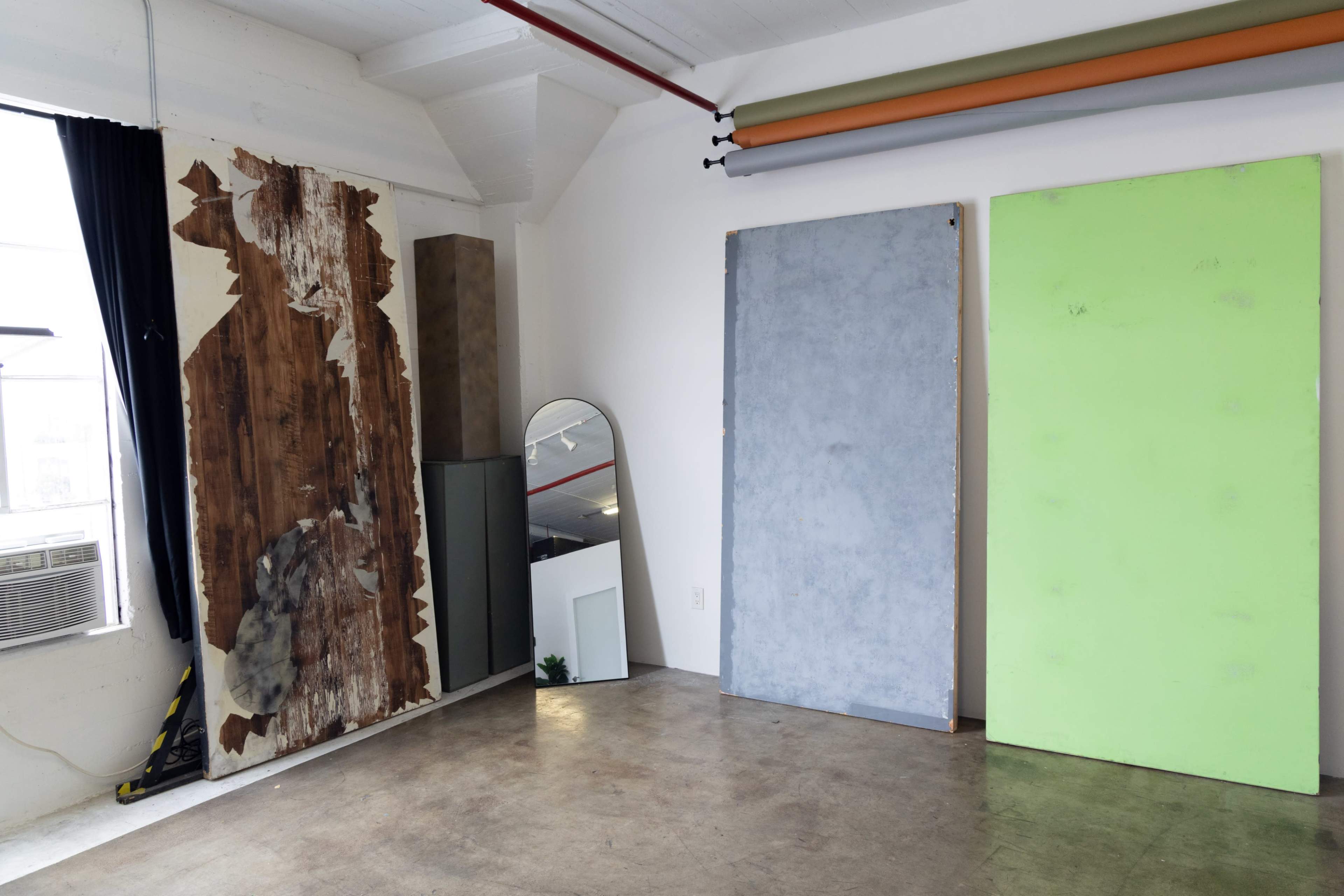 The image shows a minimalist interior space with three distinct wall panels, including a wooden texture, a gray panel, and a bright green panel, along with a mirror and an air conditioning unit.