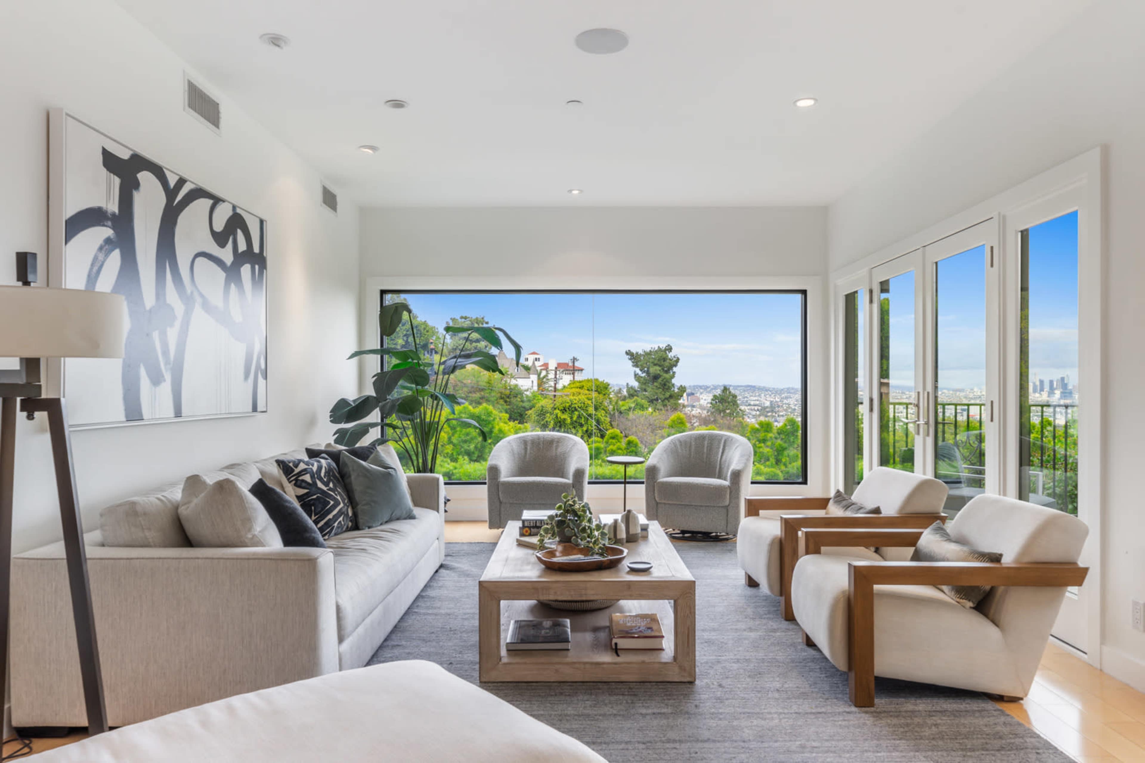 A bright living room features a large window with a view of greenery, furnished with a light-colored sofa, two armchairs, and a coffee table.