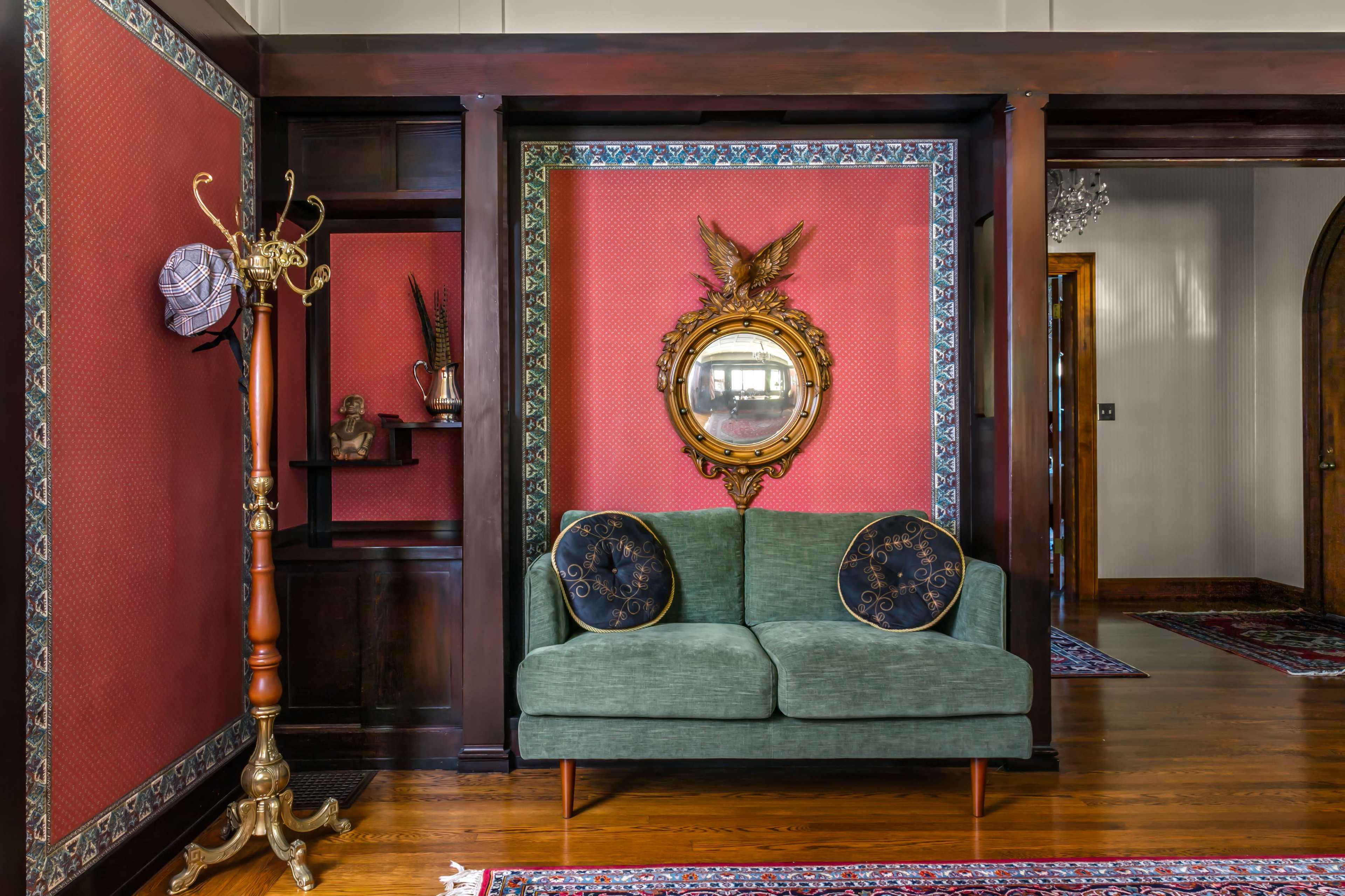 The image shows a vintage-style sitting area featuring a green two-seater sofa with decorative pillows against a red patterned wall, complemented by a round mirror and a coat stand.