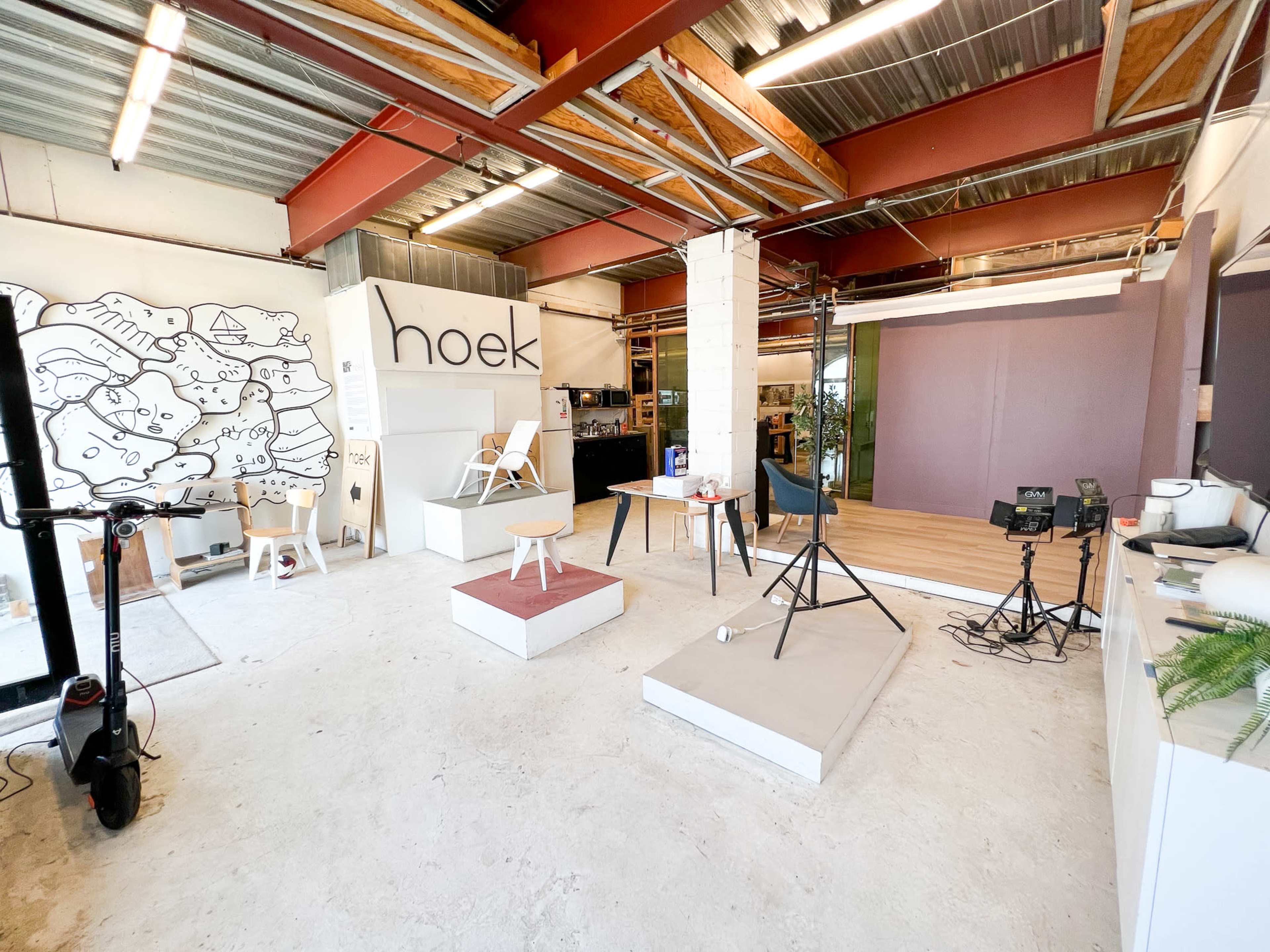 The image shows an industrial-style studio space with white walls, exposed beams, and various set areas, including a backdrop for photography and a decorative mural.