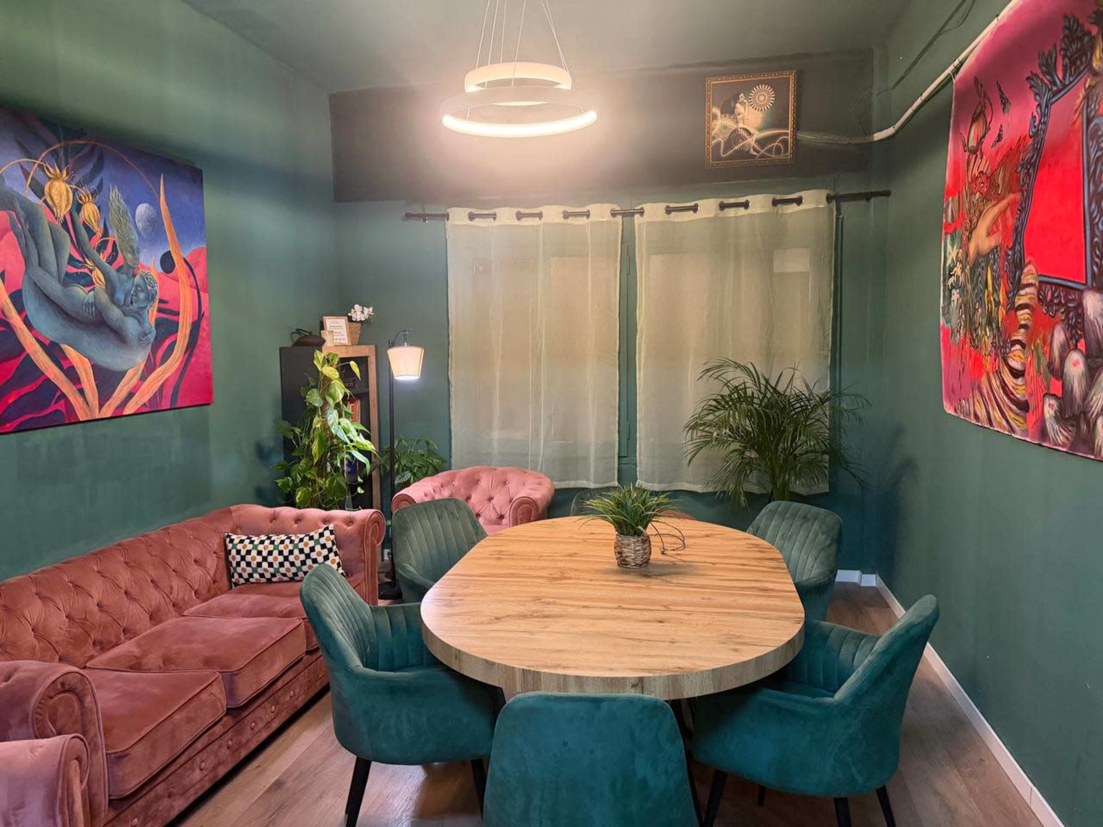 The image shows a cozy room with green walls, featuring a round wooden table surrounded by green chairs and a pink velvet couch, along with vibrant artwork on the walls.