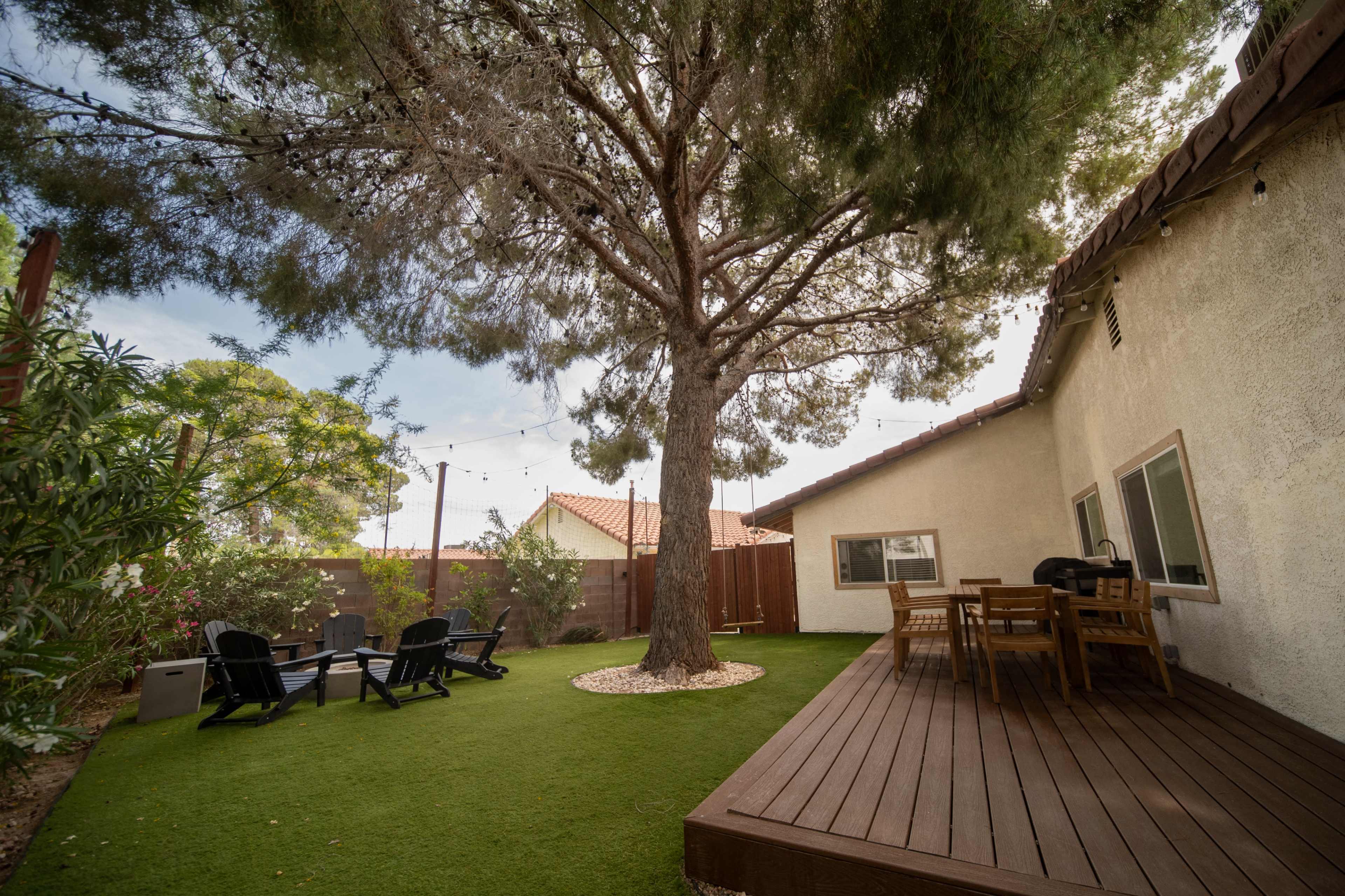 The image shows a backyard with a large tree, a wooden deck, and black patio chairs on a green lawn.