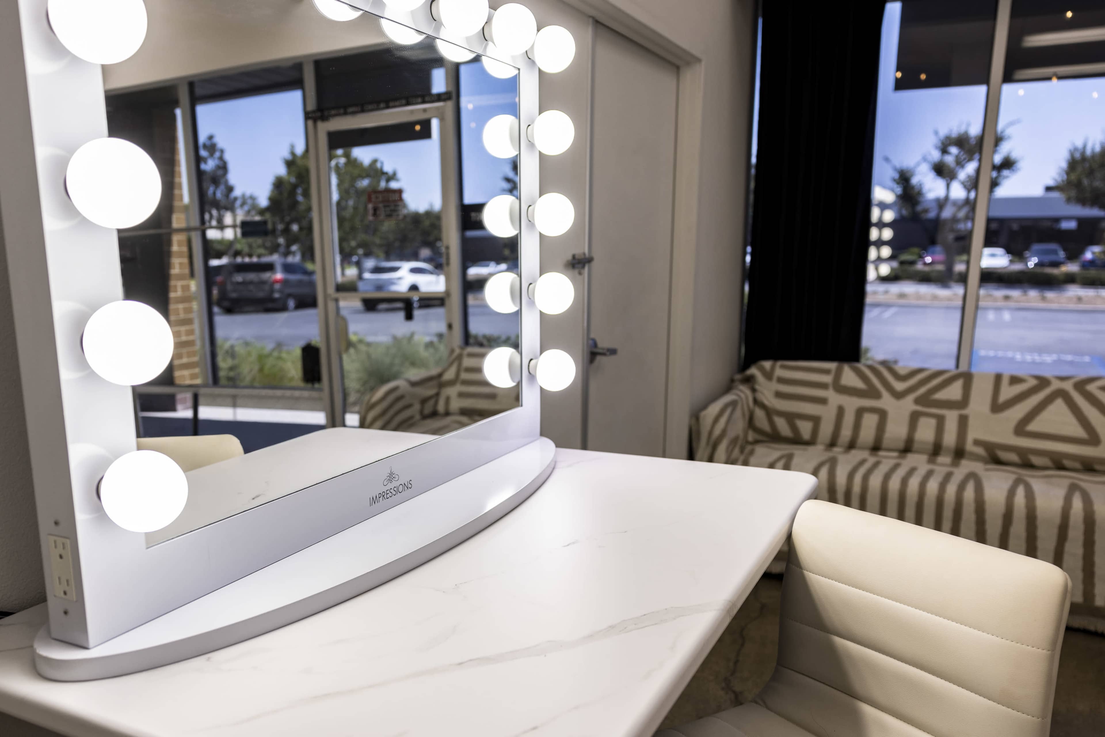 The image shows a well-lit vanity mirror with large bulbs, positioned on a white table next to a patterned couch and large windows revealing a parking lot outside.