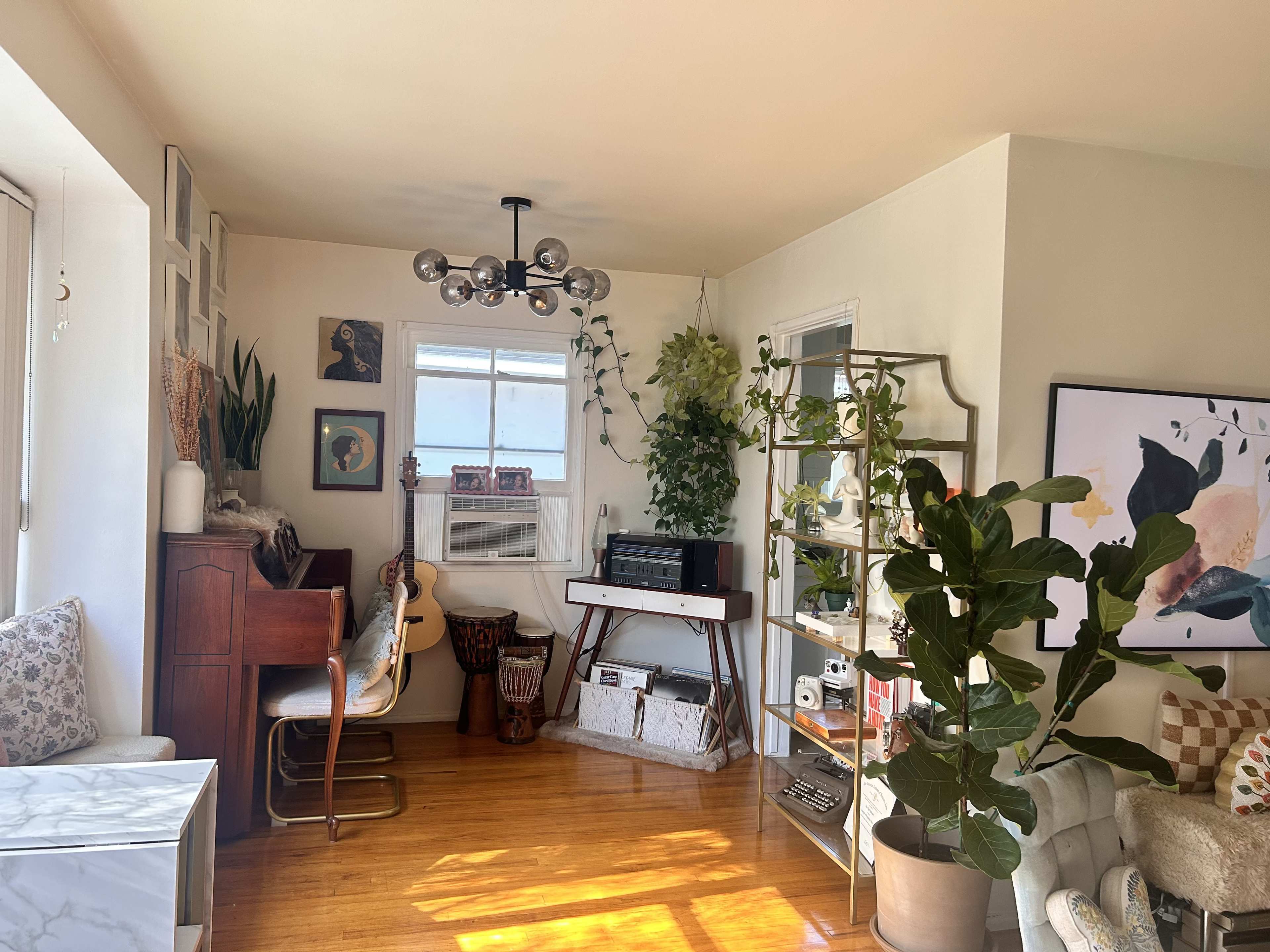 A cozy living room features a mix of plants, a wooden piano, an acoustic guitar, and a large window allowing natural light to enter.