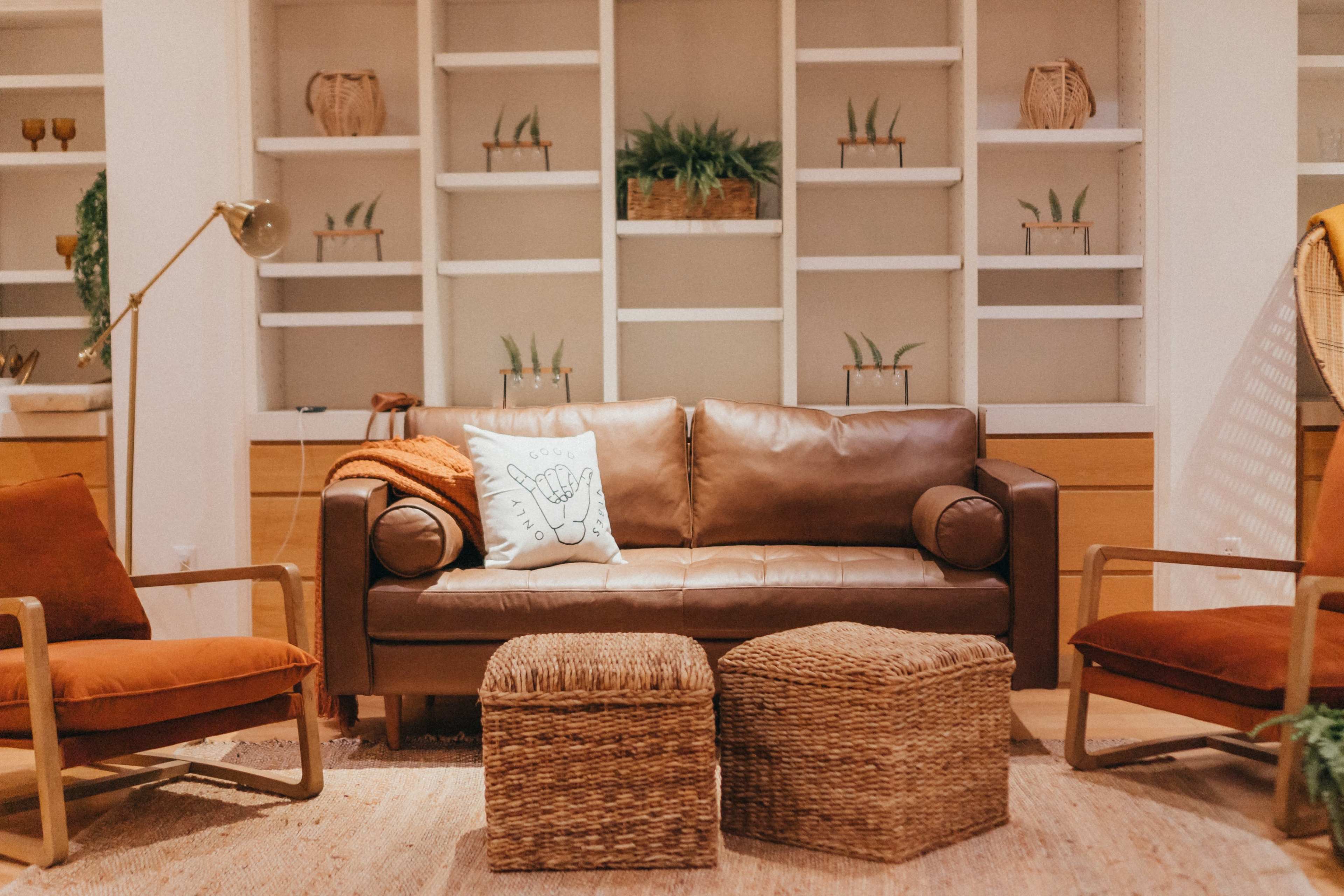 A cozy living room with a brown leather couch, two wooden chairs, and woven ottomans arranged around a soft area rug, with a shelf in the background featuring plants and decorative items.