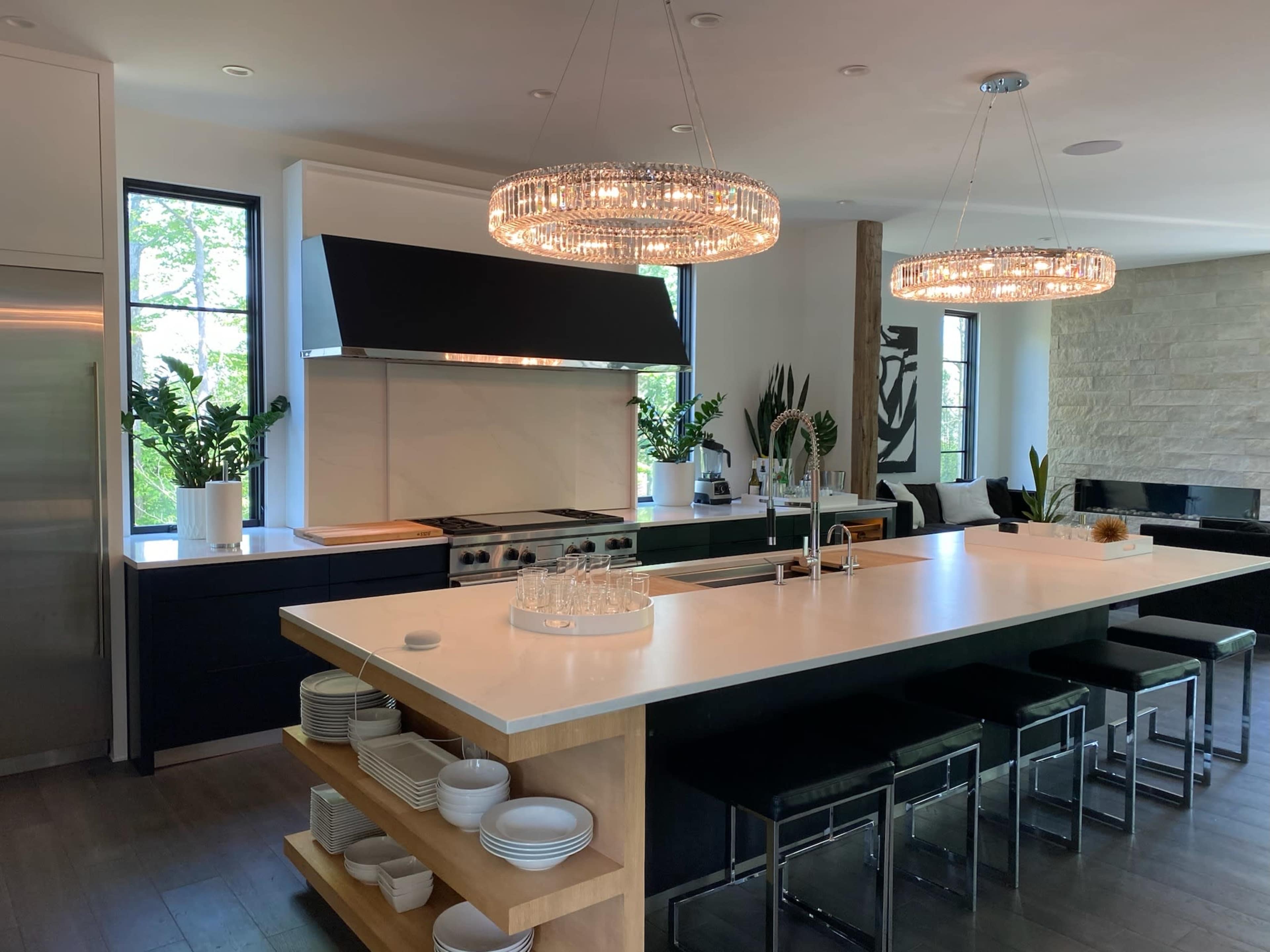 The kitchen features a large island with a white countertop, black cabinetry, and pendant lighting above, complemented by windows and greenery.