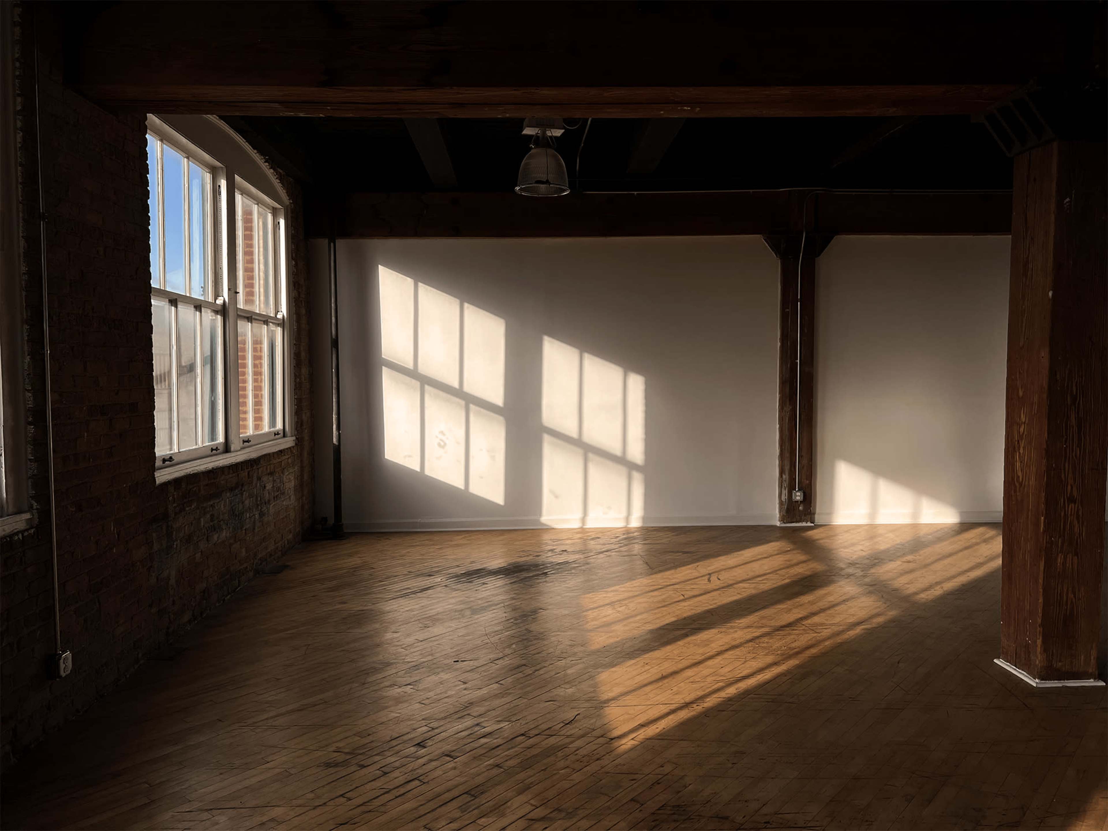 Sunlight casts shadows of large windows onto the wooden floor of an empty room with exposed brick walls and wooden beams.