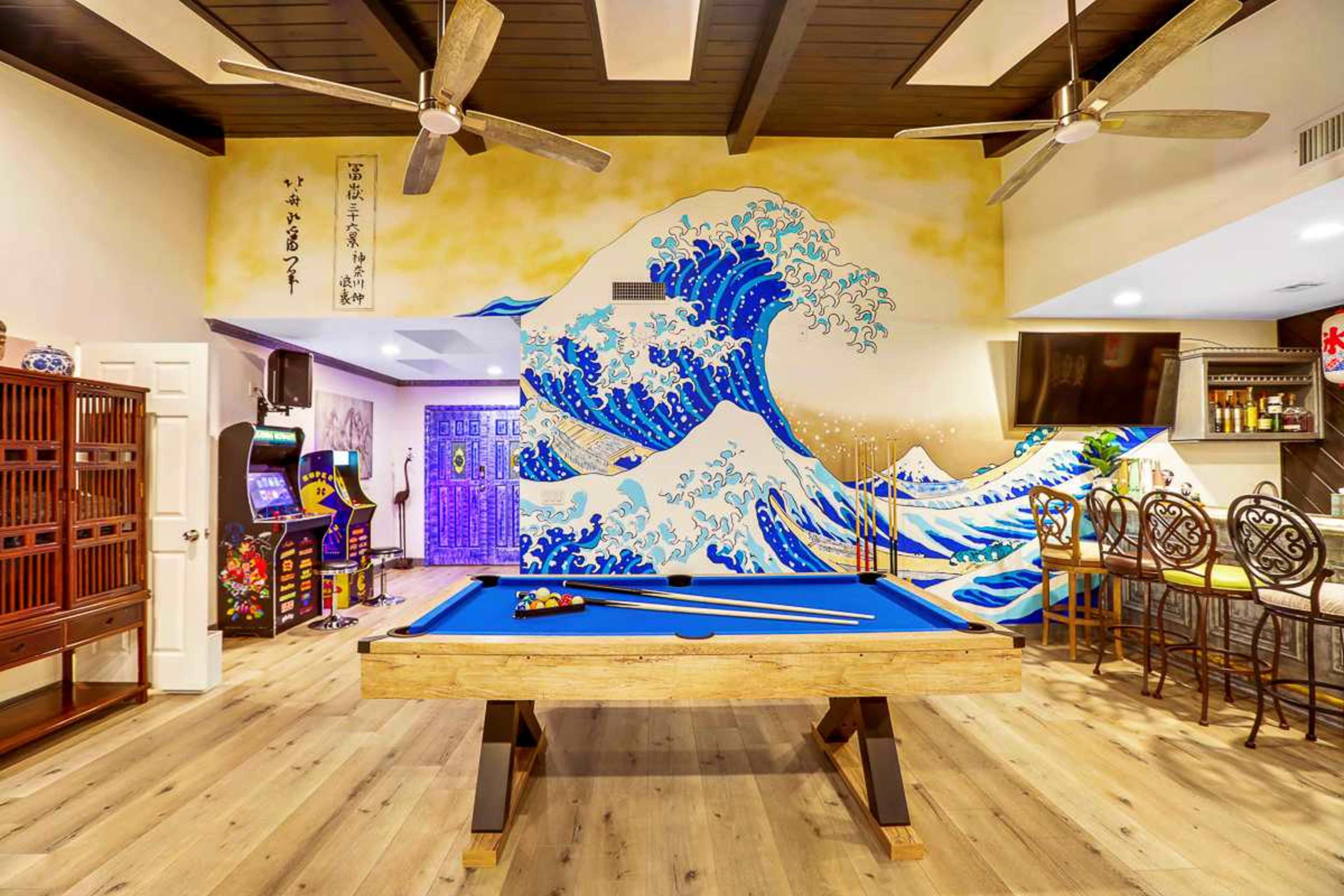 A game room with a blue pool table, a large wave mural on the wall, and various gaming machines and bar stools nearby.