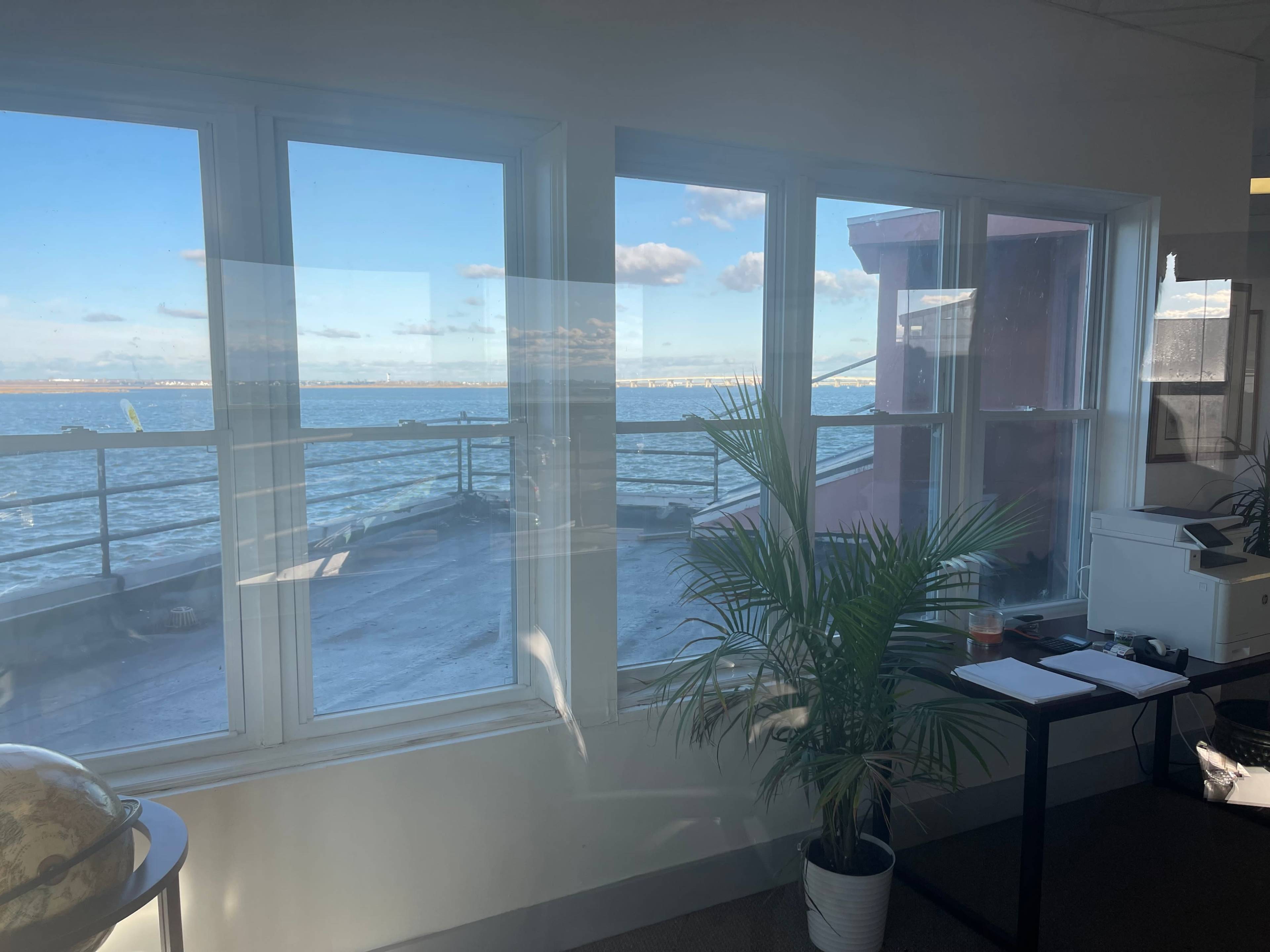 A view of the water from an office space, with large windows, a potted plant, and office furniture visible.