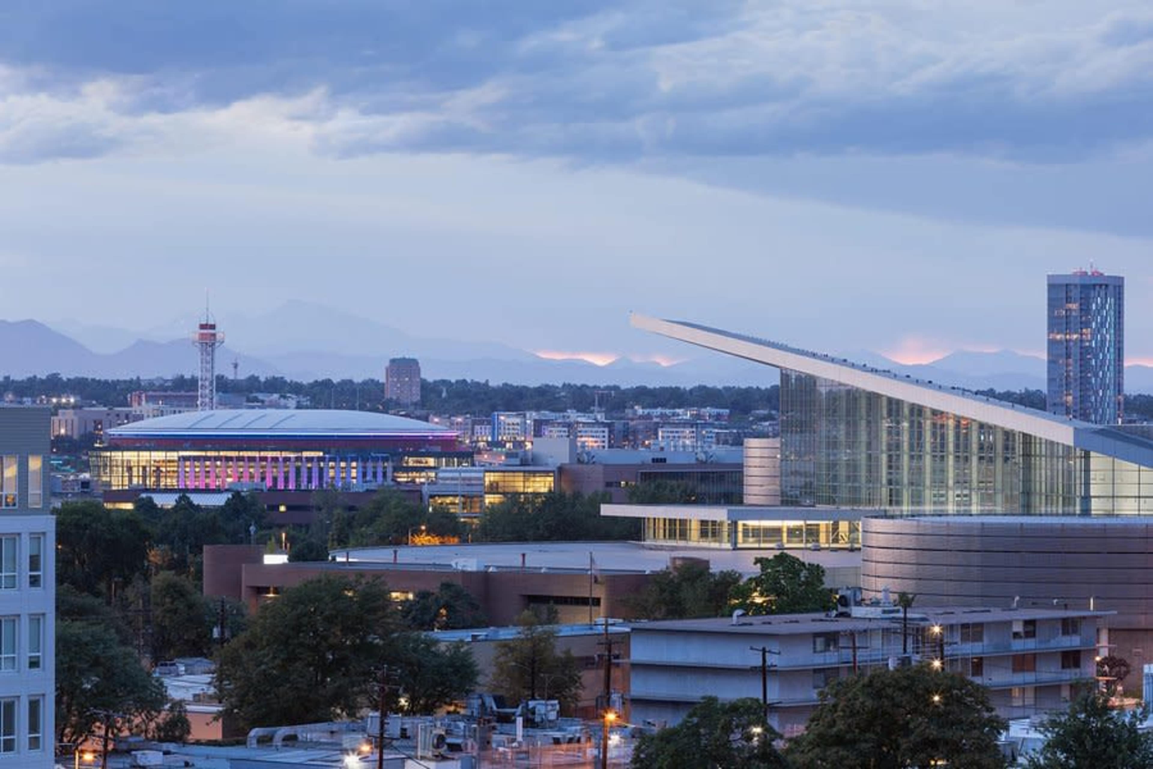 The image shows a skyline view of a city with modern buildings, including a large arena, under a cloudy sky with distant mountains in the background.