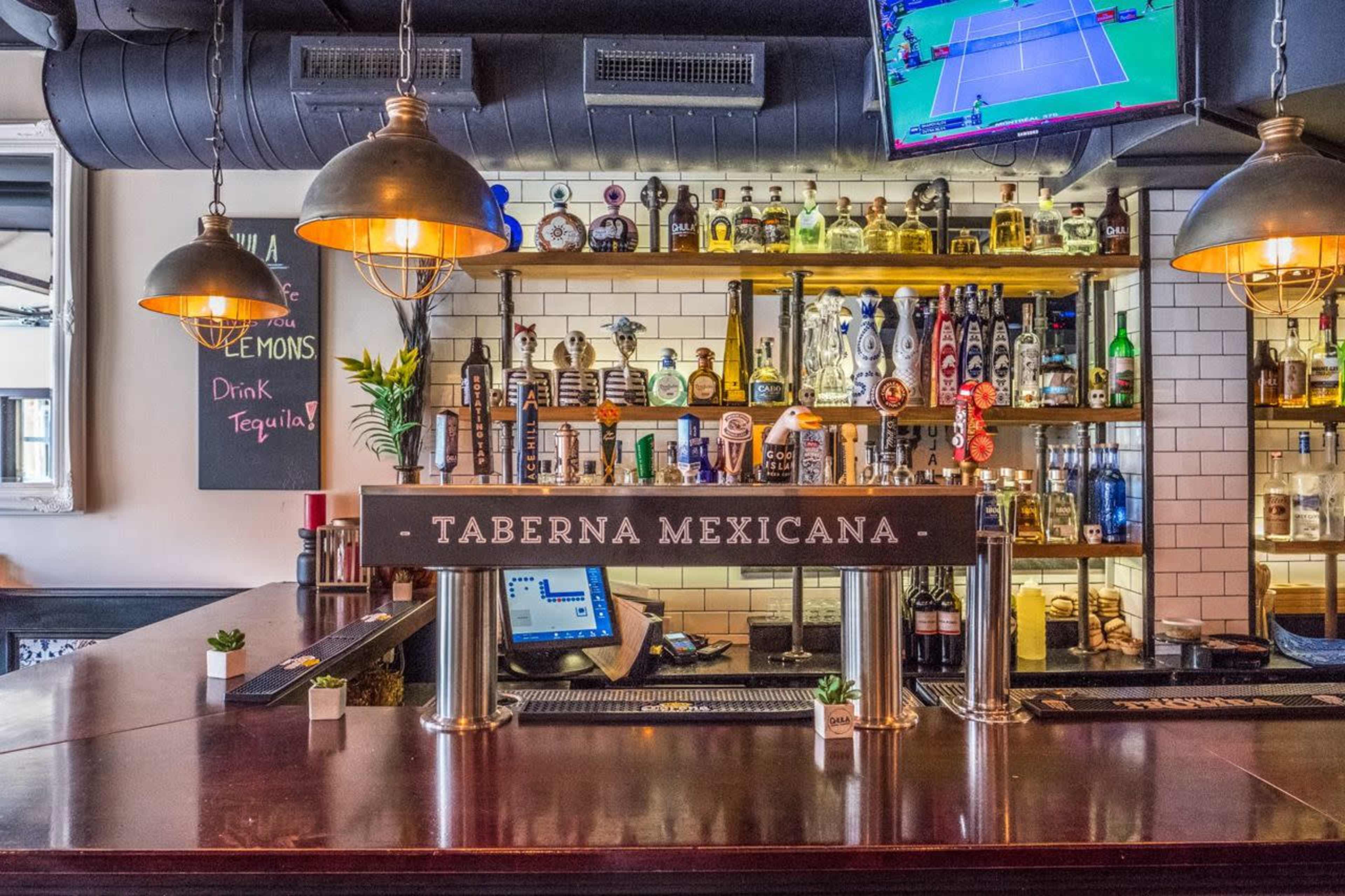The image shows a well-lit bar area with various alcohol bottles displayed on shelves, a large bar counter, and a illuminated sign reading "TABERNA MEXICANA."