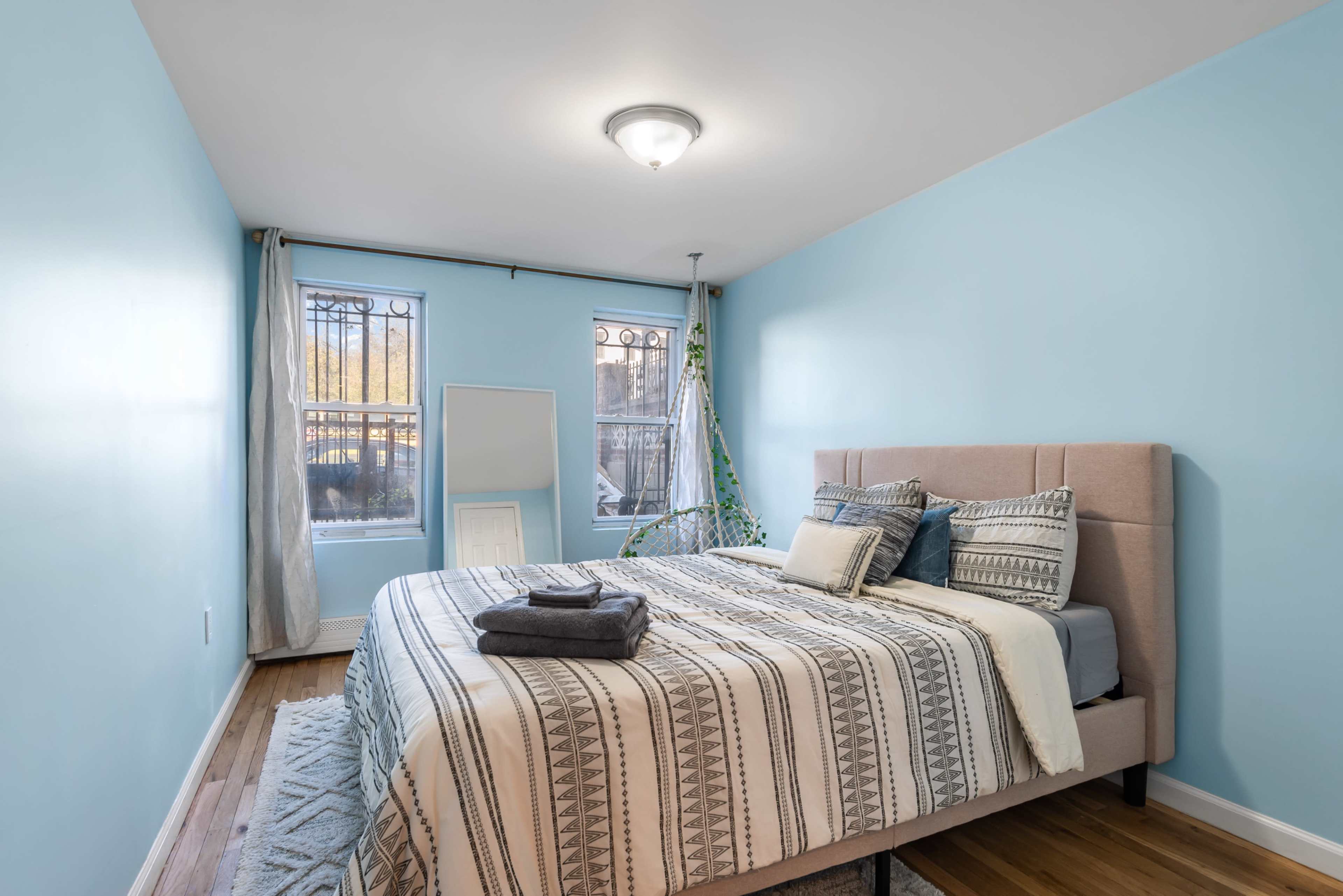 A neatly arranged bedroom features a light blue wall, a bed with patterned bedding, and two windows with sheer curtains.