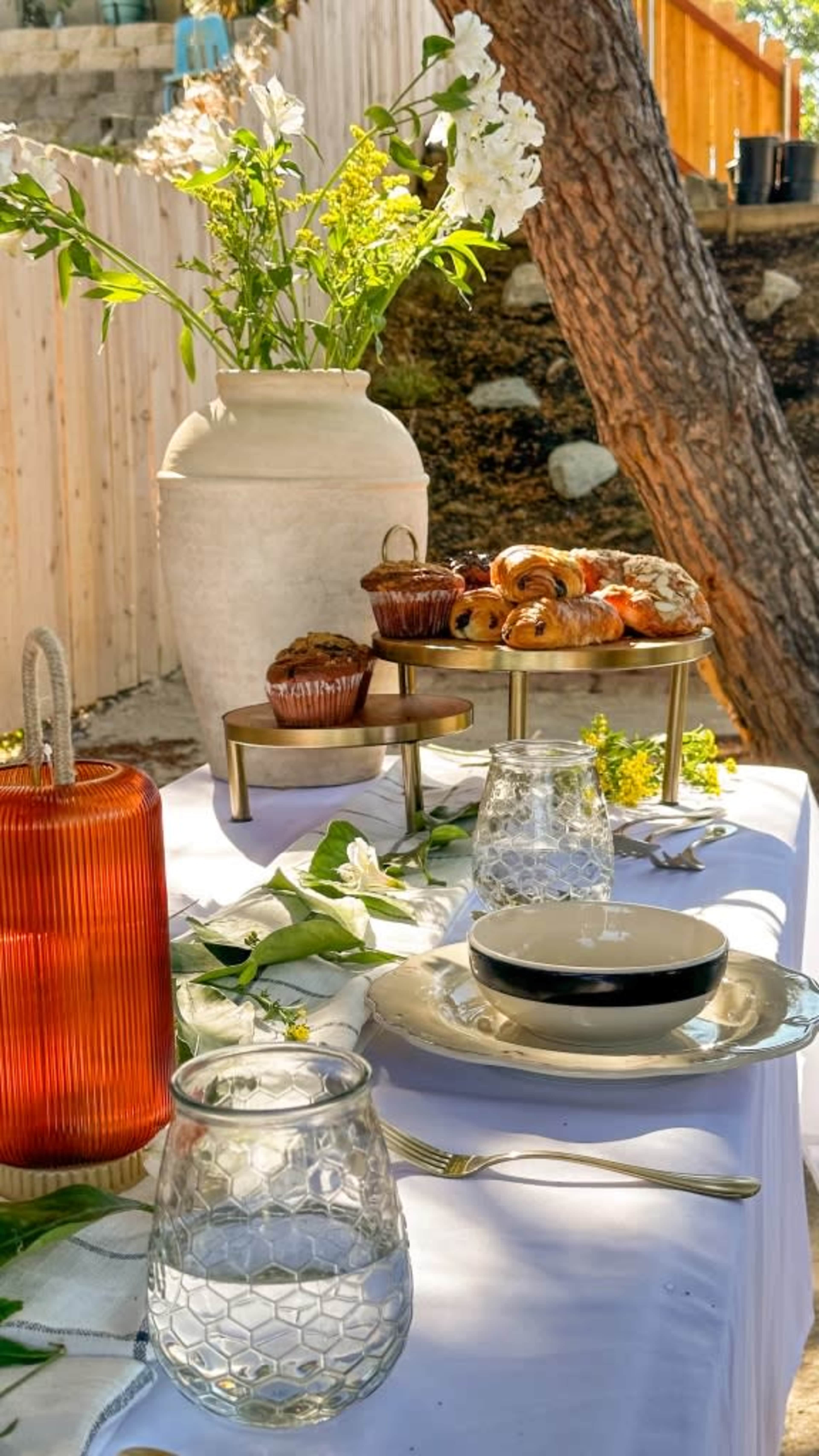 A elegantly set outdoor table displays a tiered stand of baked goods alongside decorative glassware and a large vase of flowers.