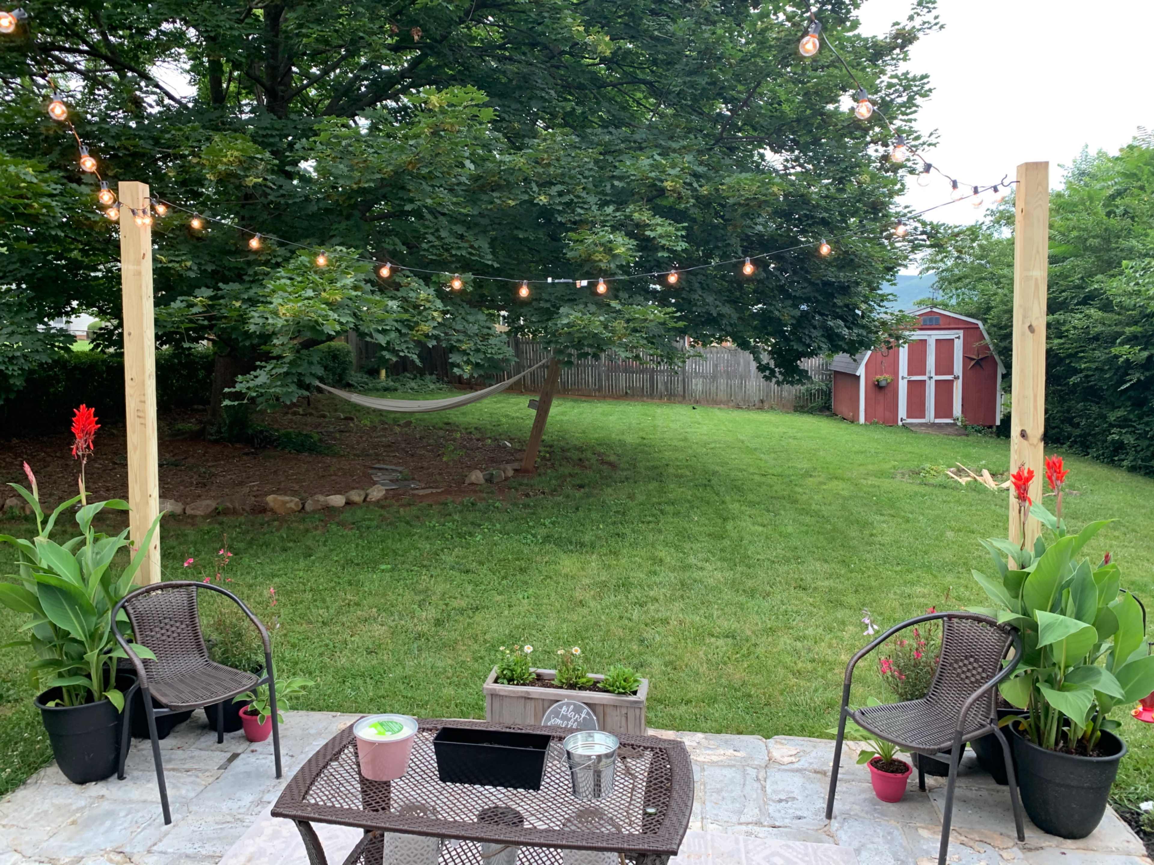 A backyard scene with two chairs and a table on a stone patio, surrounded by greenery and string lights overhead, with a hammock hanging nearby and a shed visible in the background.