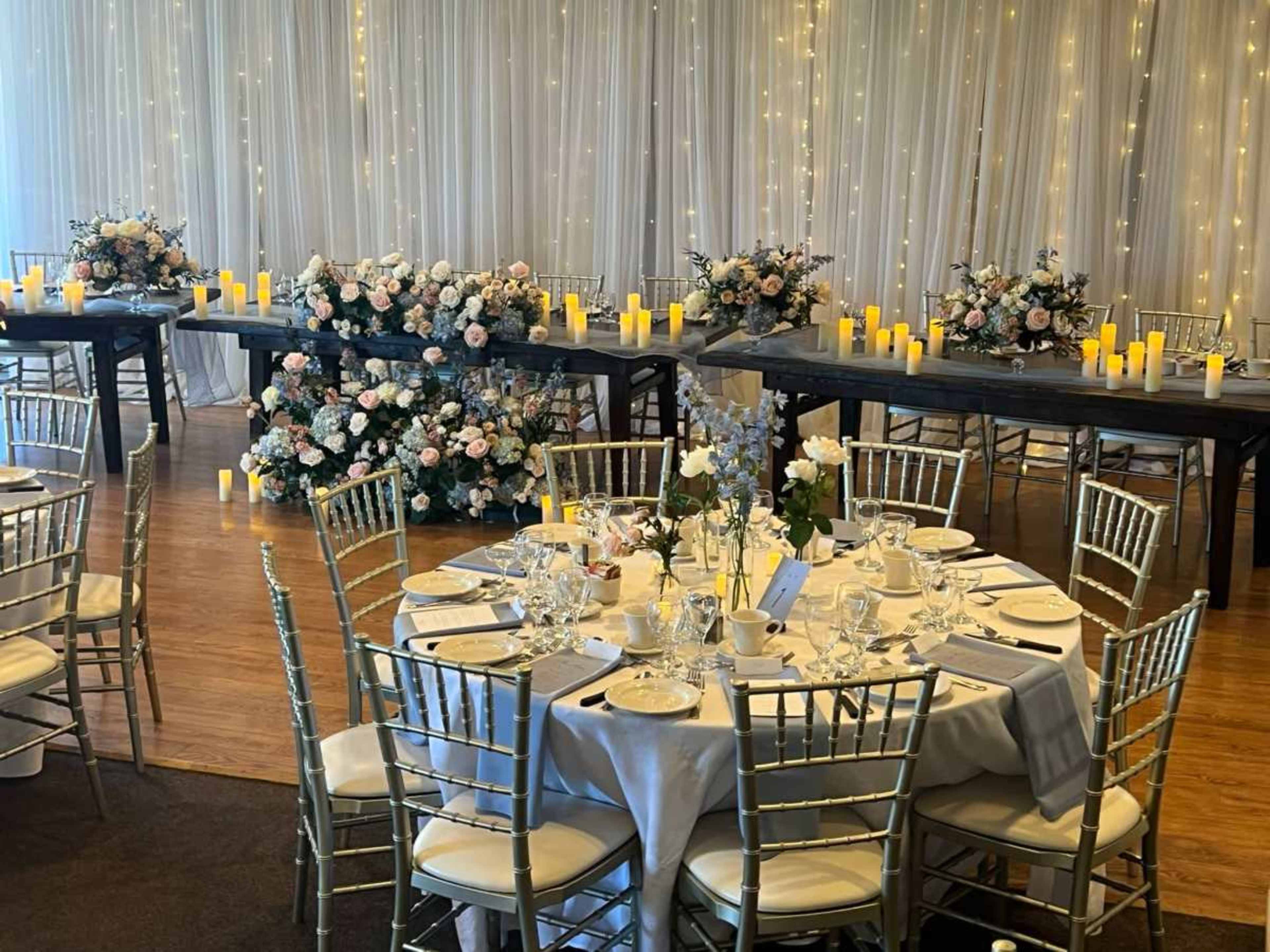 The scene features elegantly set round tables with silver chairs in a decorated venue, showcasing floral arrangements and lit candles against a backdrop of draped fabric.