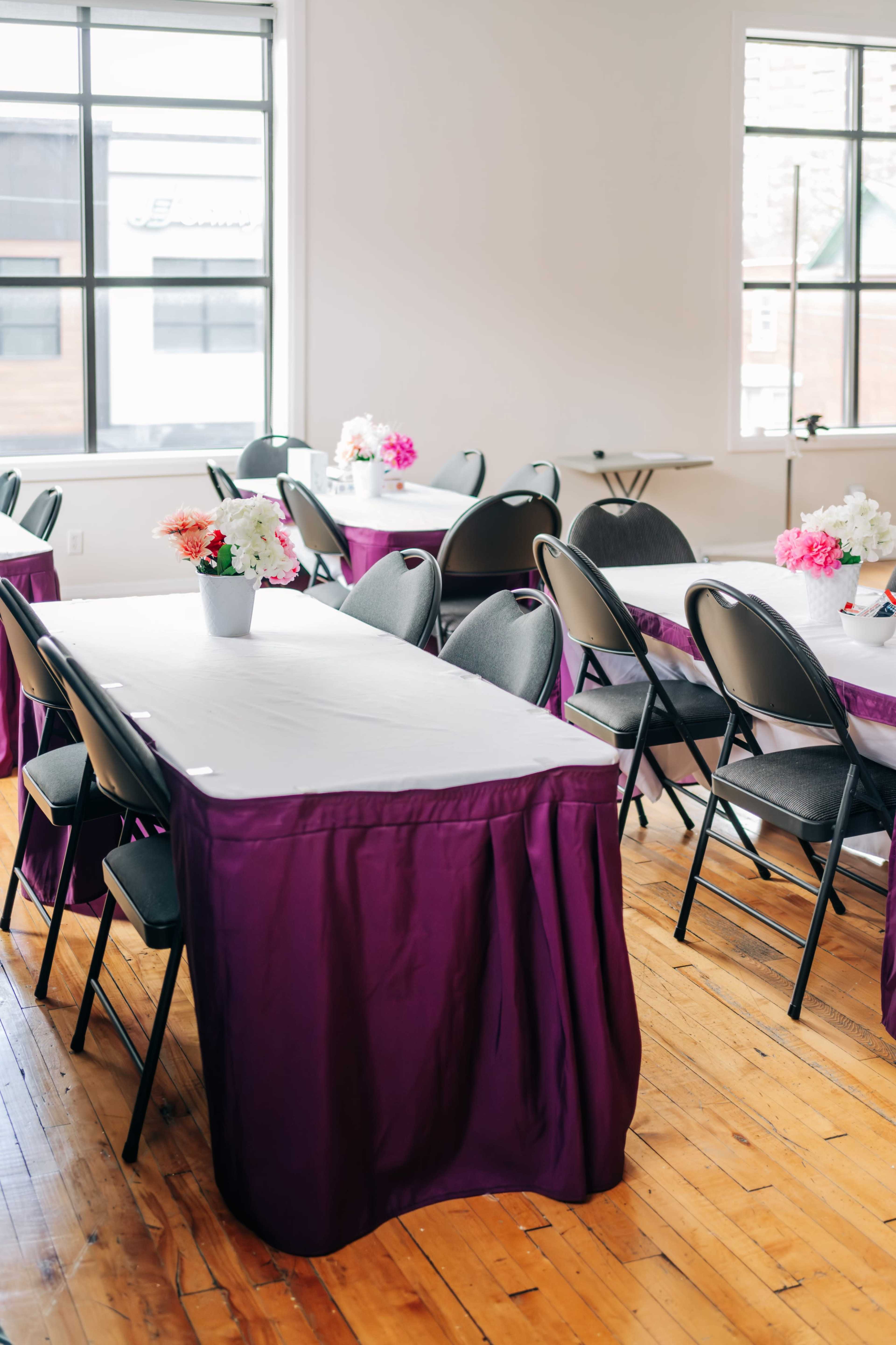 A spacious room features several tables covered with purple tablecloths and adorned with floral centerpieces, surrounded by black chairs.