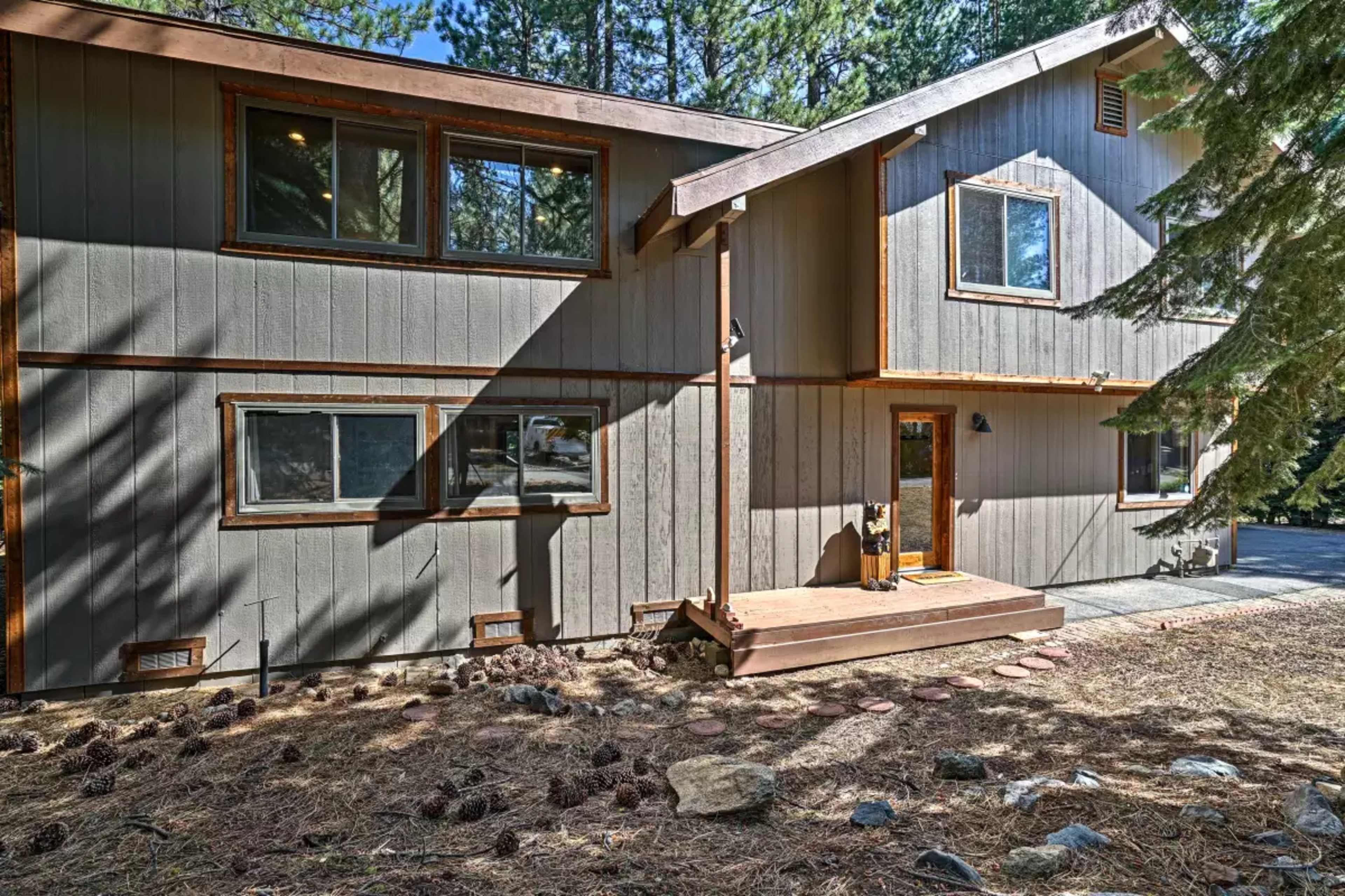 A two-story wooden house with a light gray exterior, surrounded by pine trees and a gravel pathway.