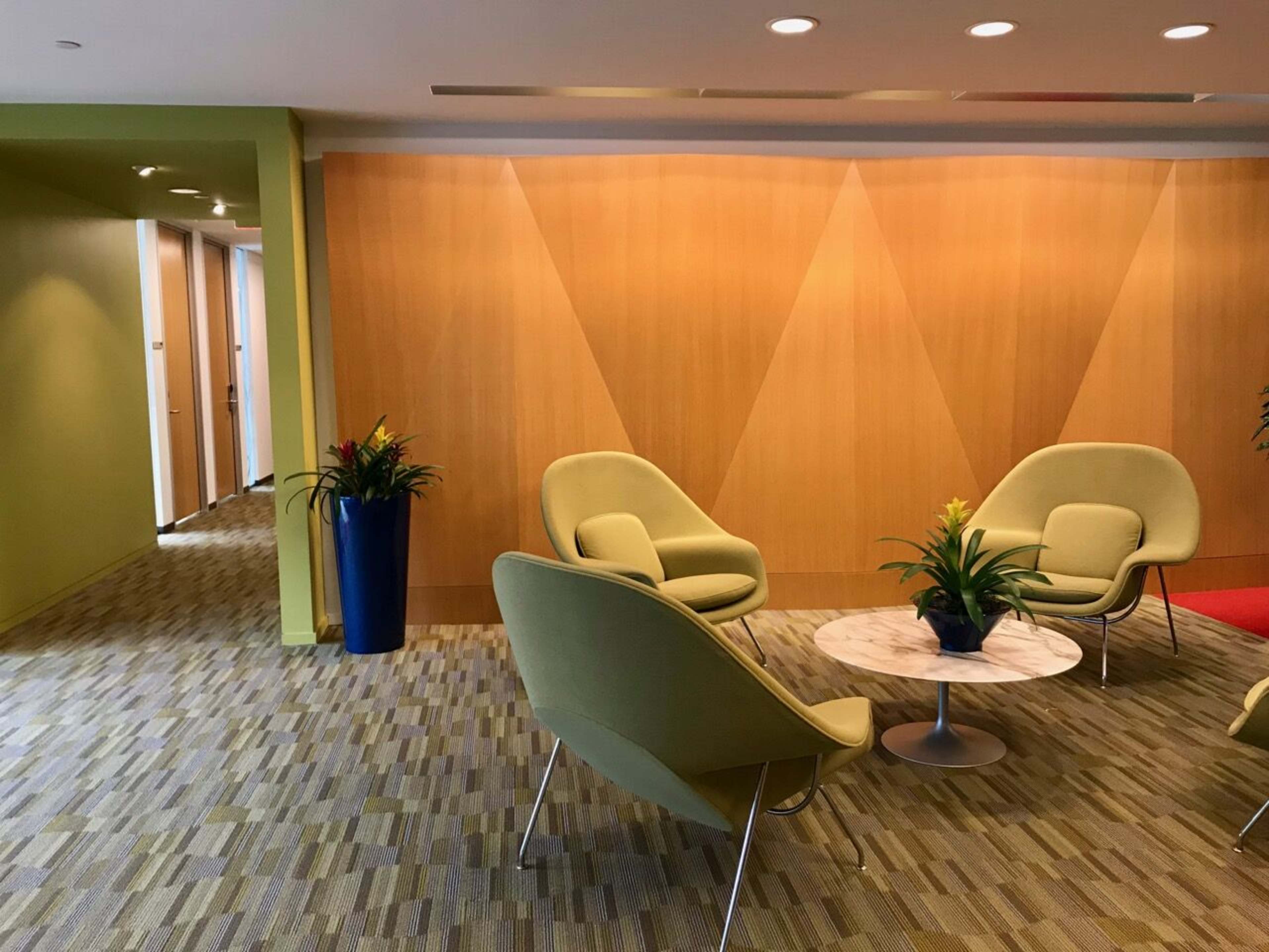 The image shows a modern waiting area featuring three green chairs, a circular table, and a tall planter with plants, set against a wooden accent wall.