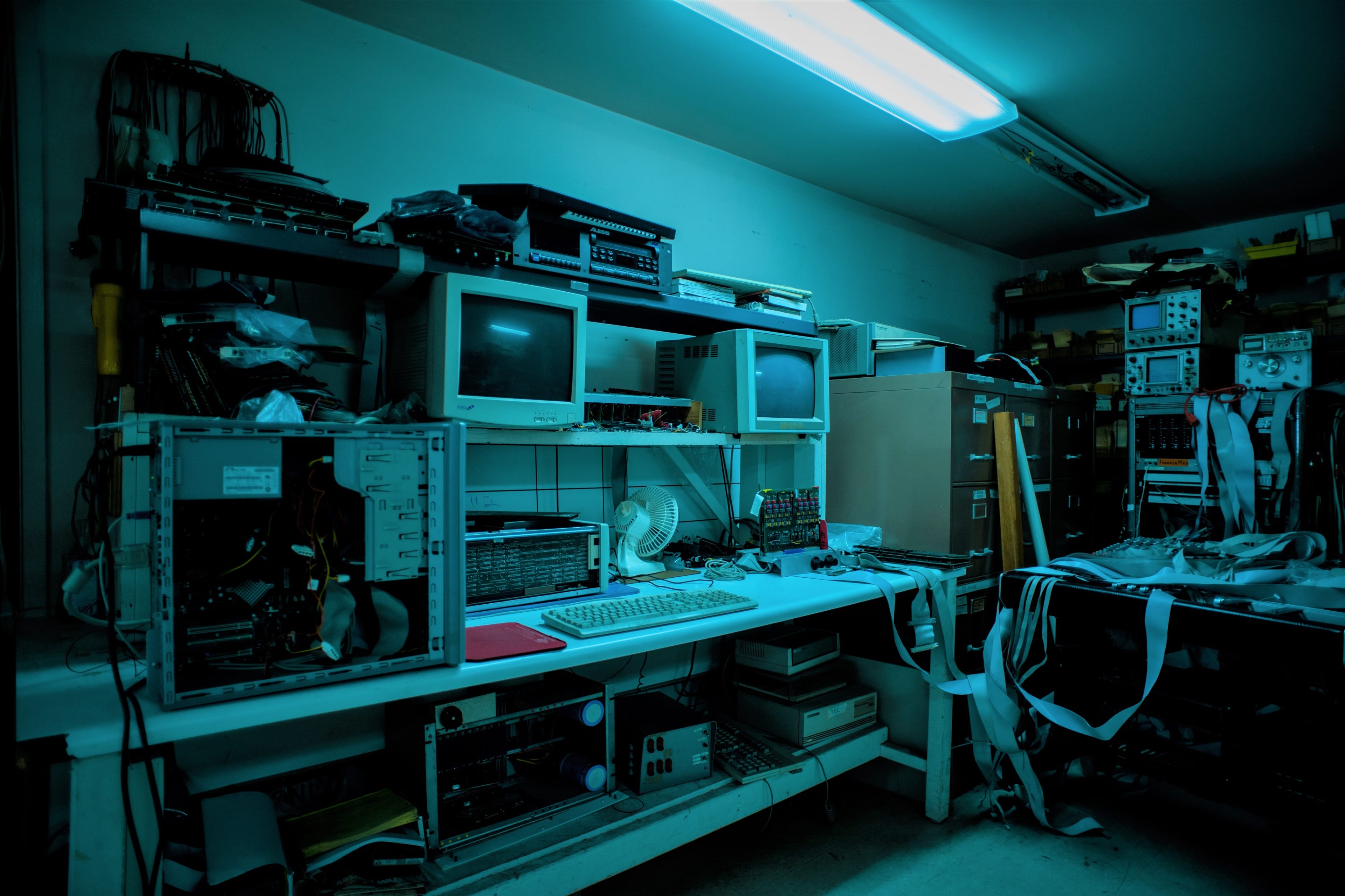 A cluttered room filled with various old computer equipment and electronics on work tables and shelves.