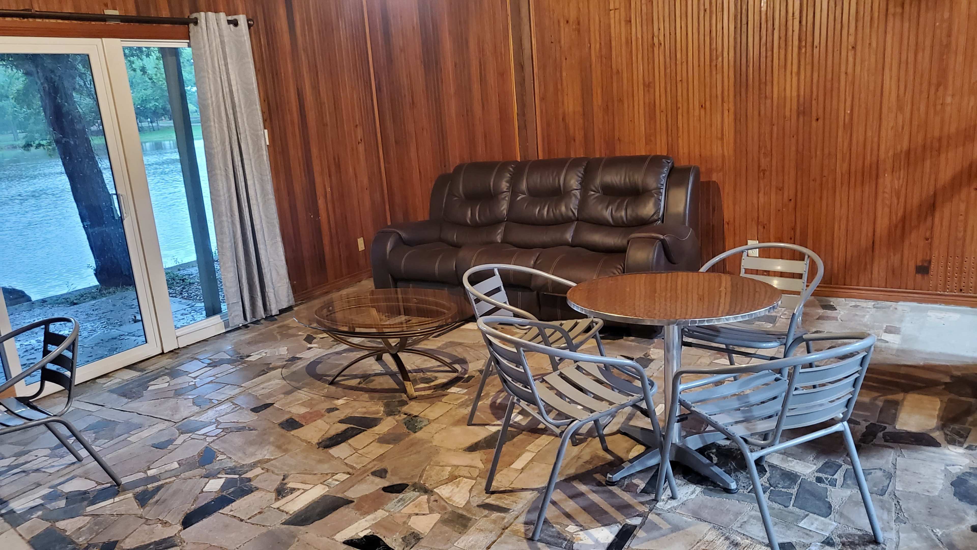 A brown leather couch and metal patio furniture are arranged in a room with wooden walls and a view of a lake through sliding glass doors.