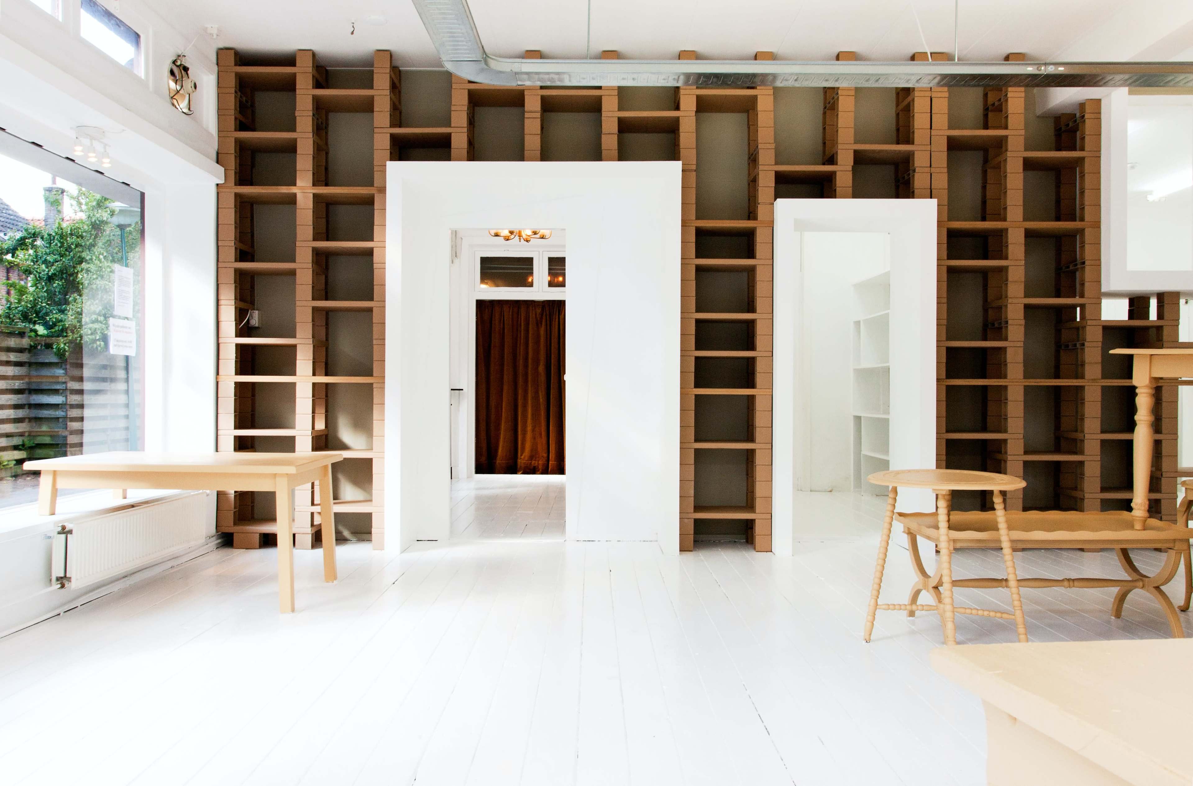 The image shows a brightly lit interior space with white floors, minimalist wooden furniture, and a wall featuring open shelving.