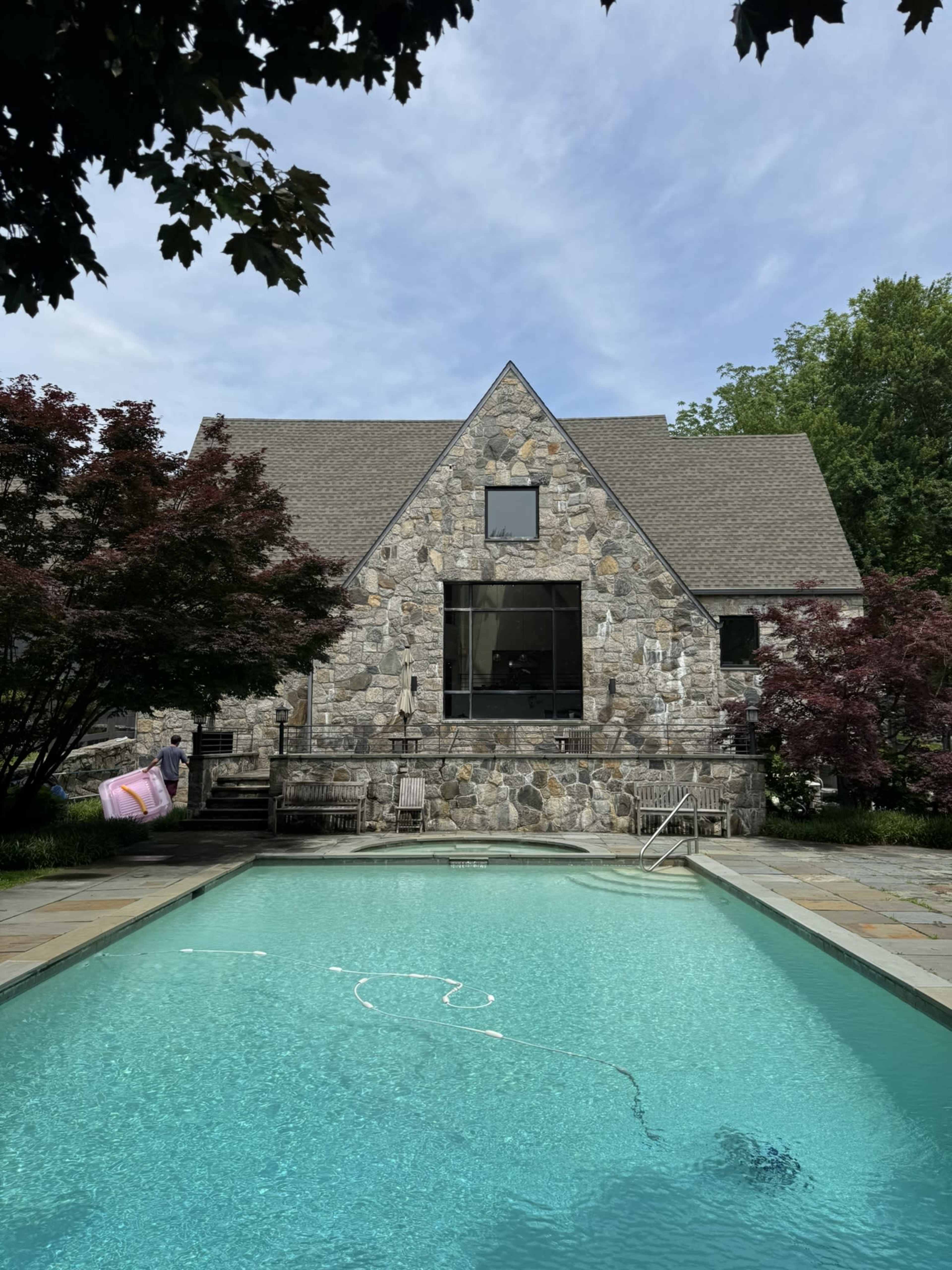A large stone house with an A-frame roof is situated beside a swimming pool, surrounded by trees and landscaping.