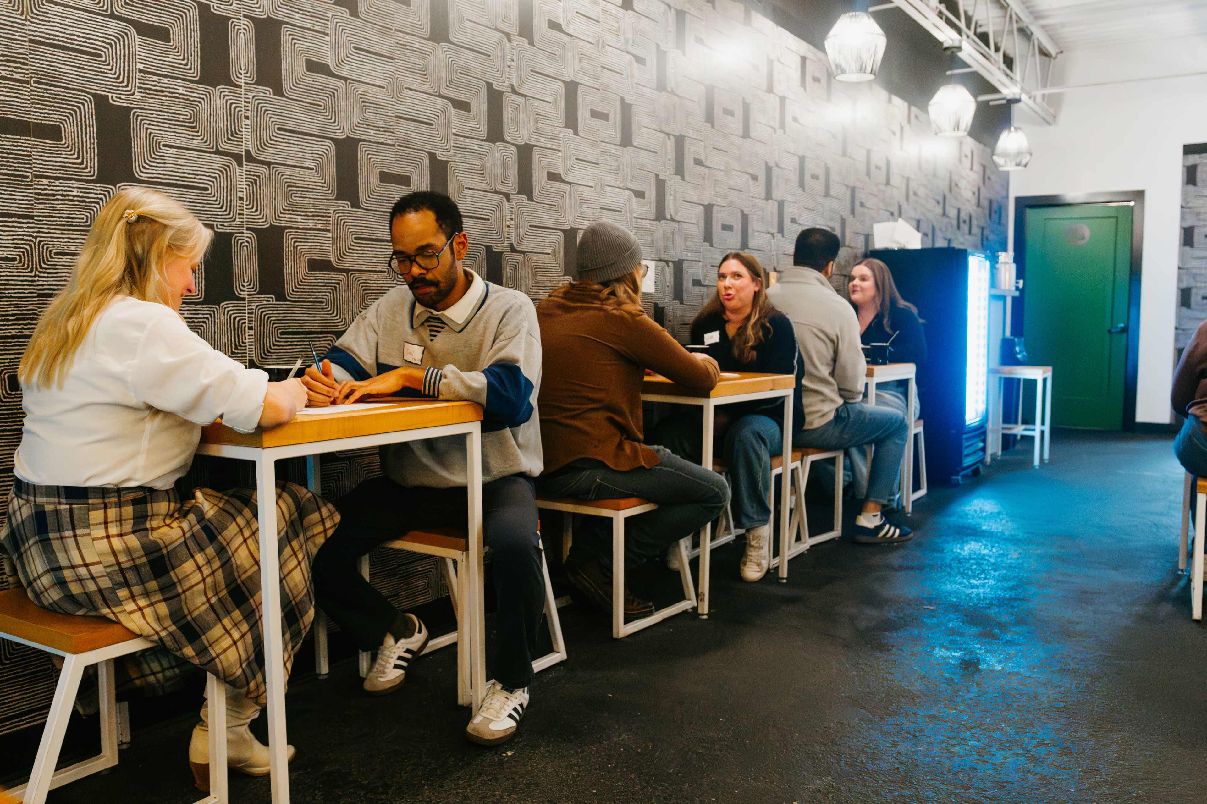 Two groups of people sit at small tables along a patterned wall in a brightly lit café.