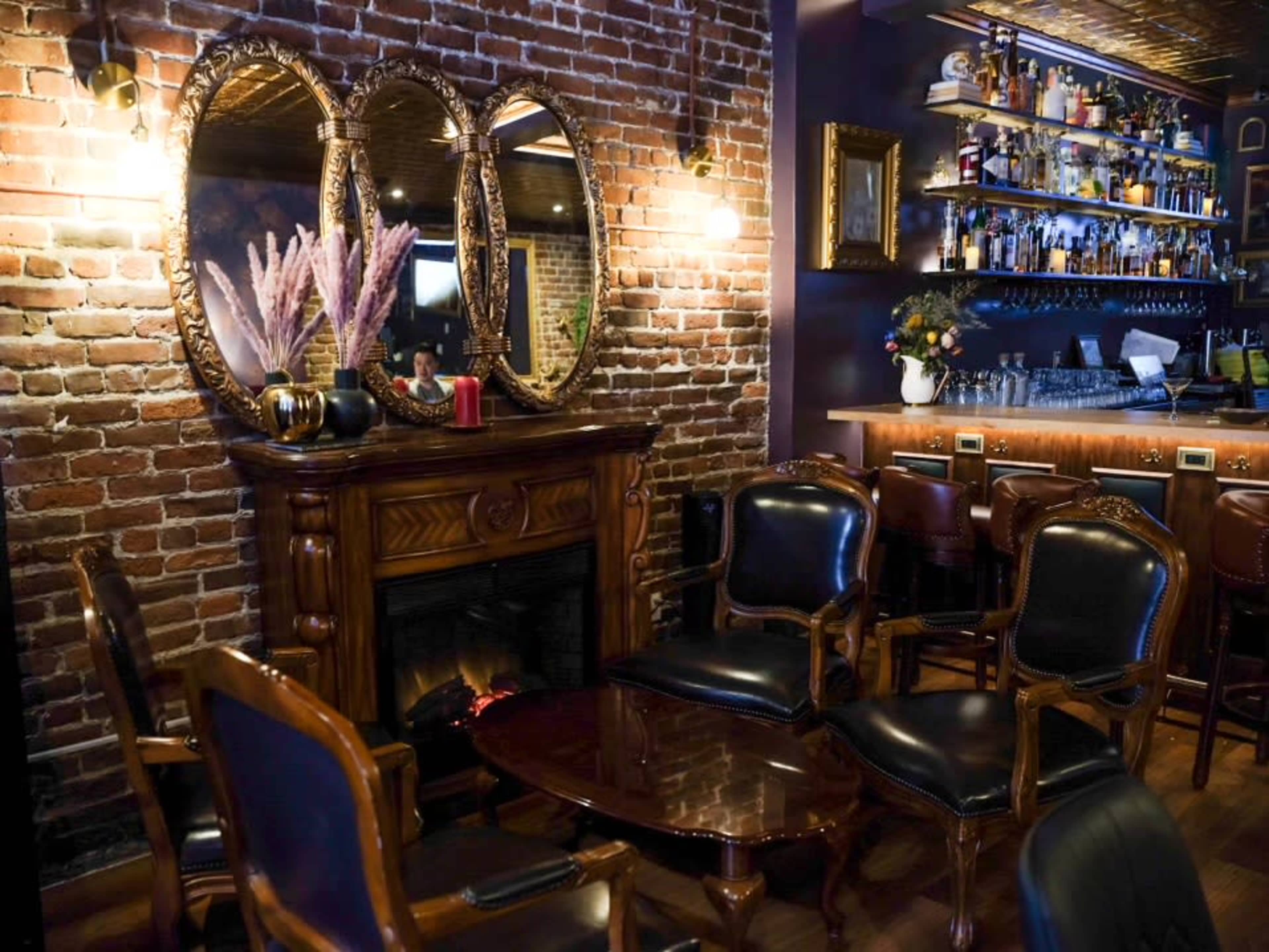 The image shows a cozy bar area with a brick wall, elegant mirrors, a wooden fireplace, and seating around a small table.