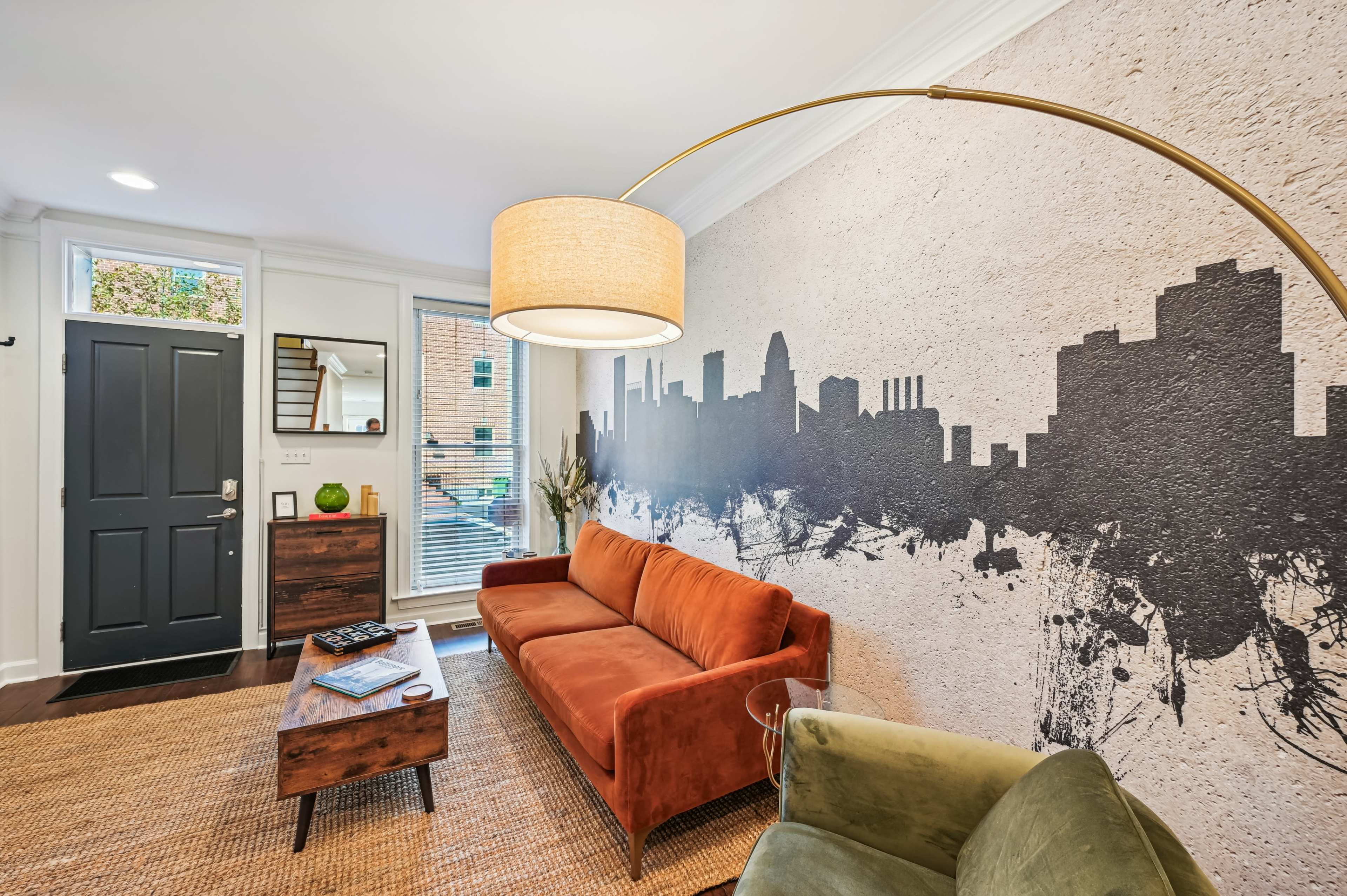 A living room features an orange sofa, a green chair, and a large wall mural of a city skyline.