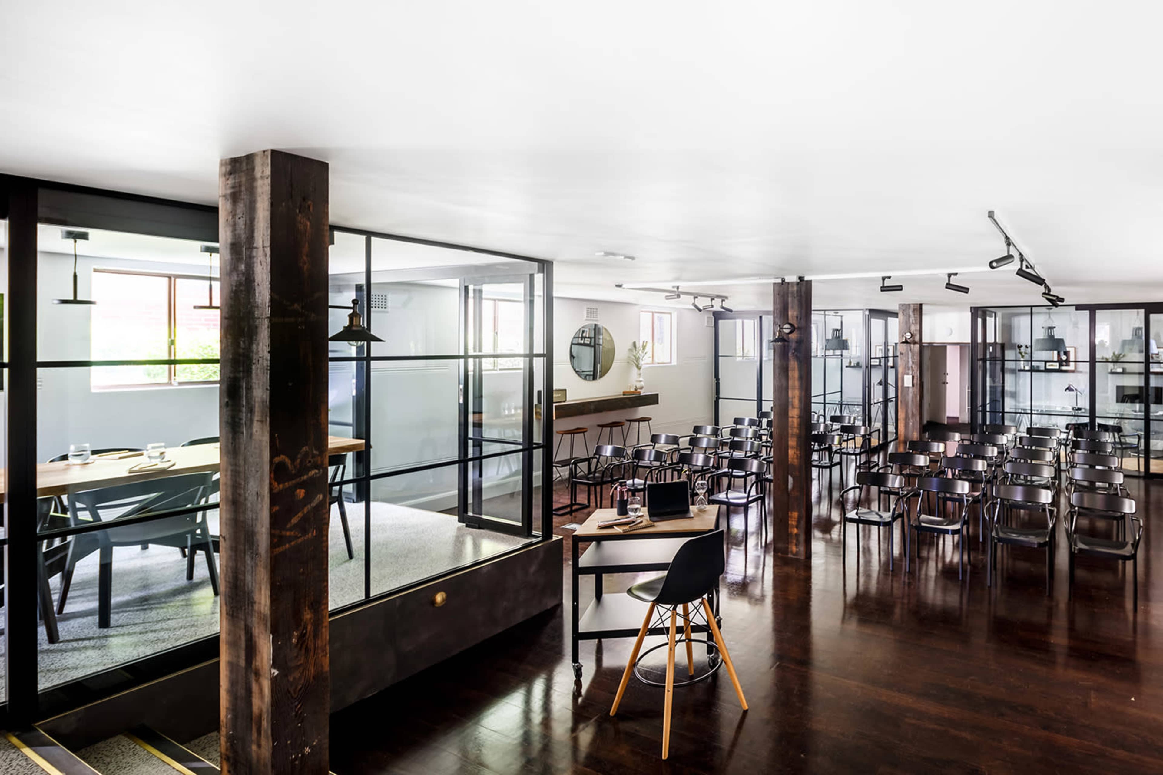 The image shows a modern meeting space with glass partitions, wooden beams, and black chairs arranged for a presentation.