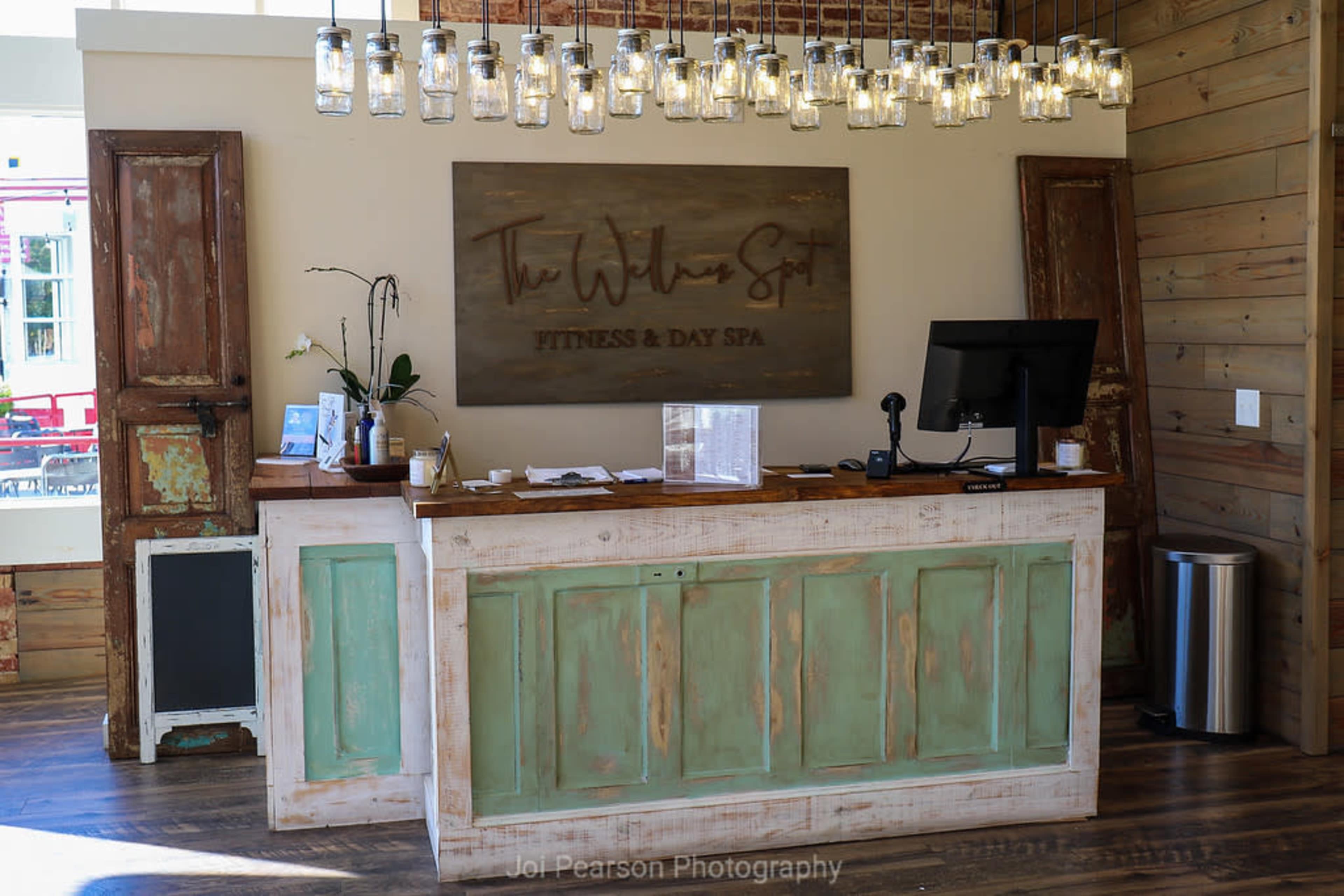 A reception desk at a spa, featuring a rustic design with green cabinetry, a large wooden sign, and hanging light fixtures.