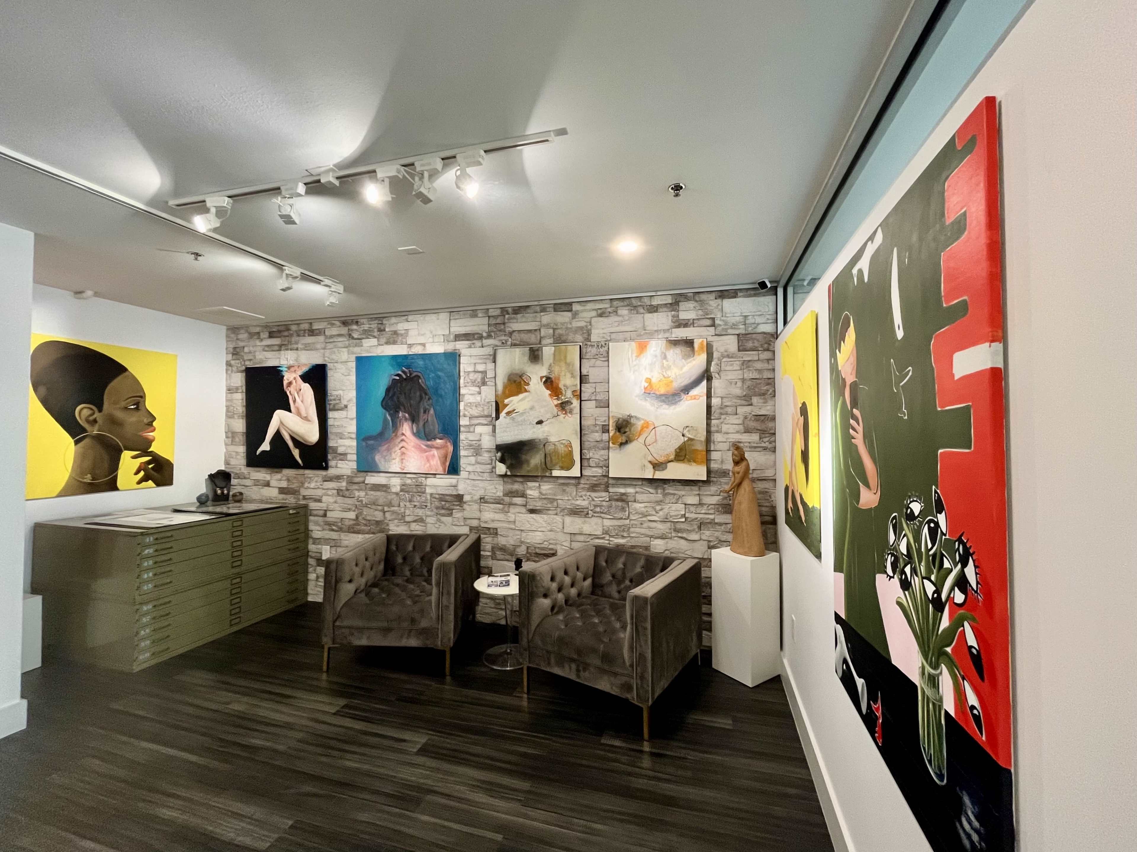 An art gallery featuring various paintings on the walls, with two upholstered chairs and a sculpture in the corner.
