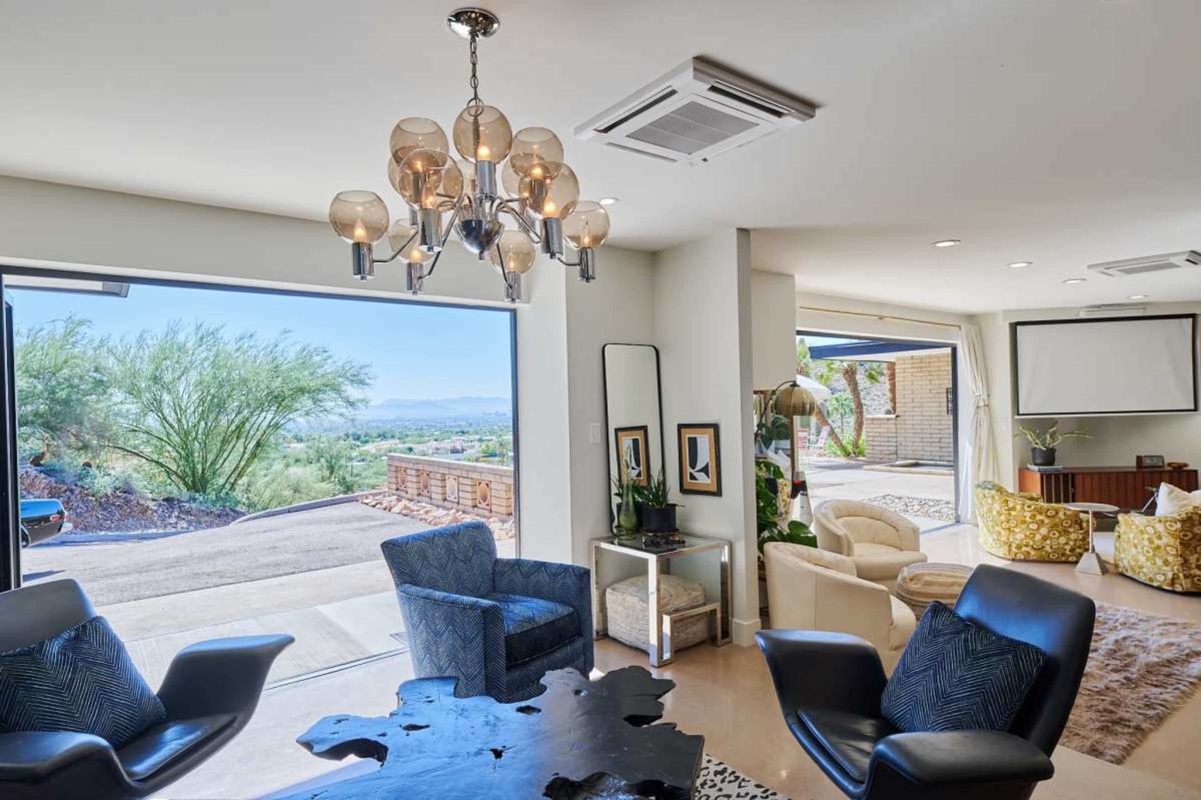 The image shows a modern living space with a chandelier, black chairs, and large glass doors that open to a scenic outdoor area.