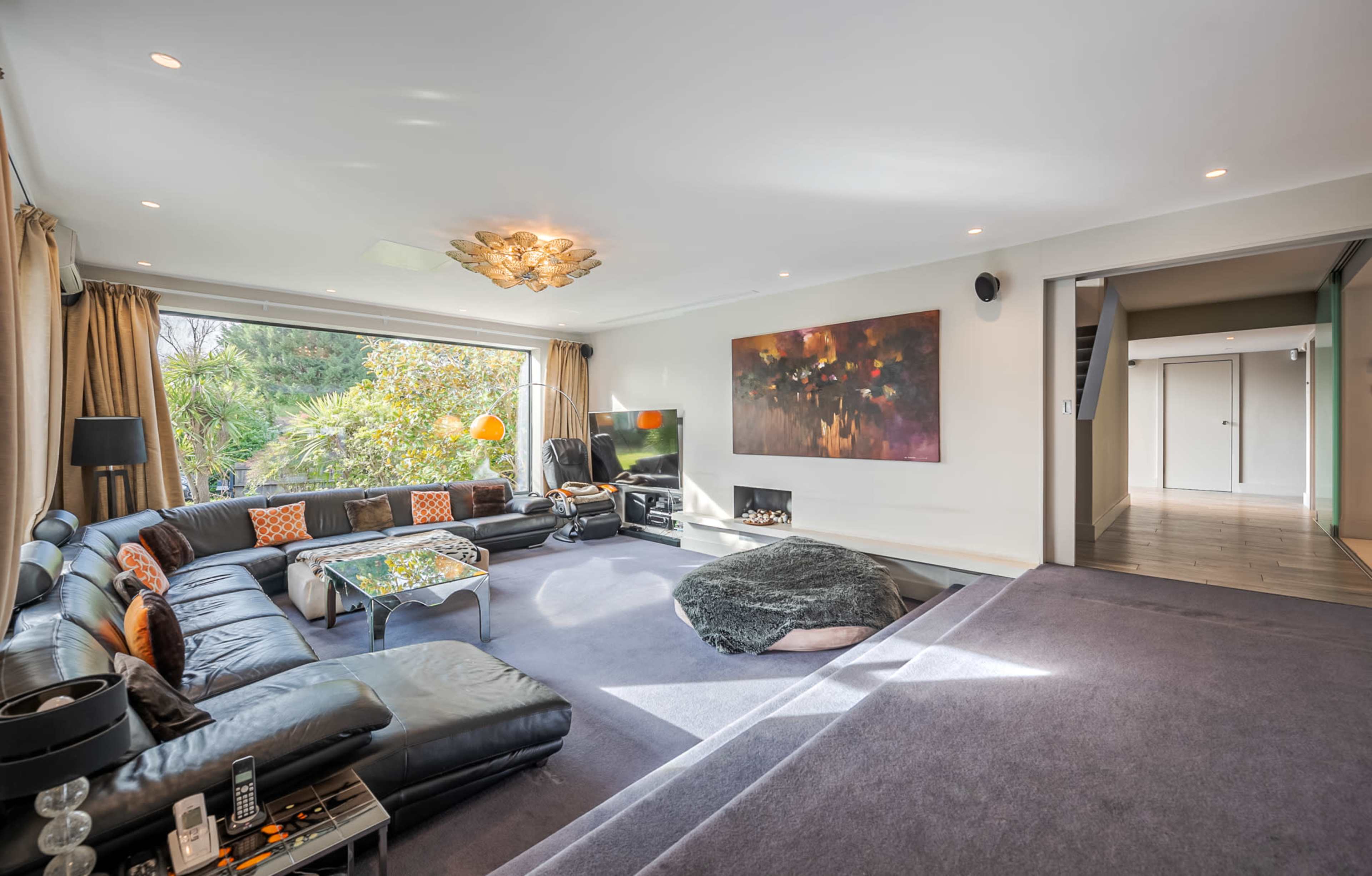 The living room features a large, sectional black sofa, a coffee table, a television, and a large window with greenery outside.