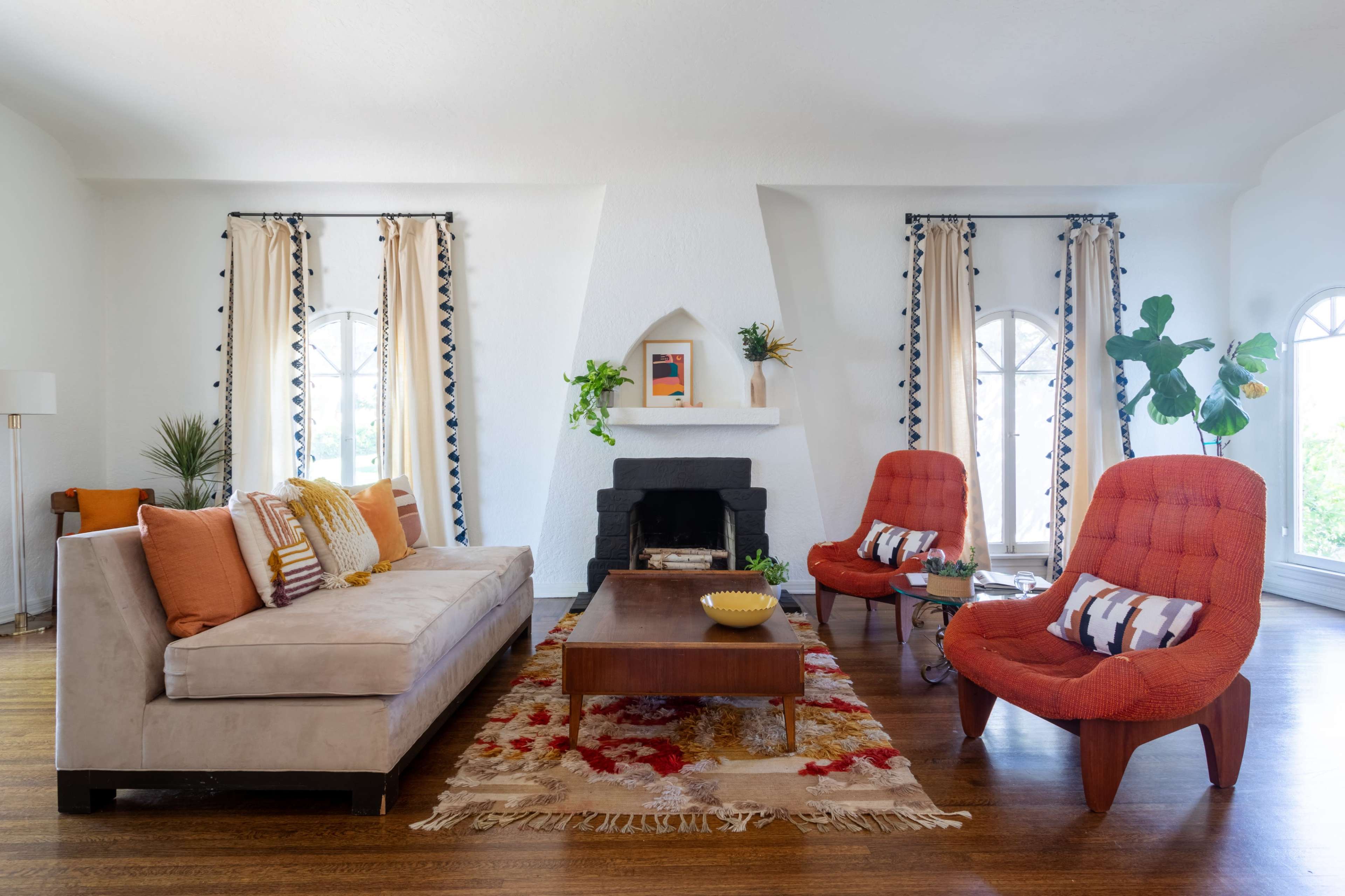 The living room features a beige sofa, two orange chairs, a wooden coffee table, and large windows with decorative curtains.