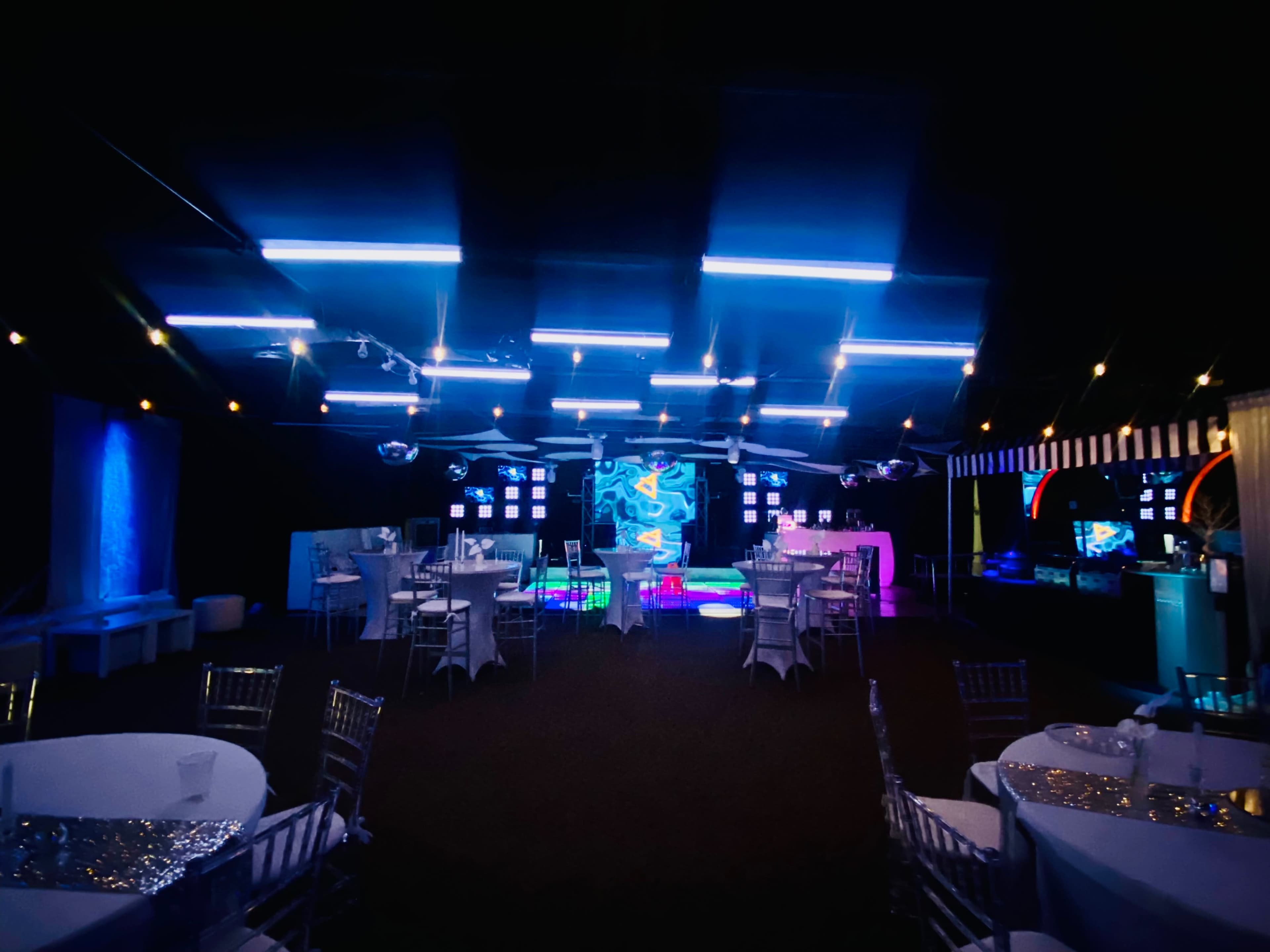 The image shows a dimly lit event space with tables and chairs arranged around a colorful backdrop featuring neon lights and screens.