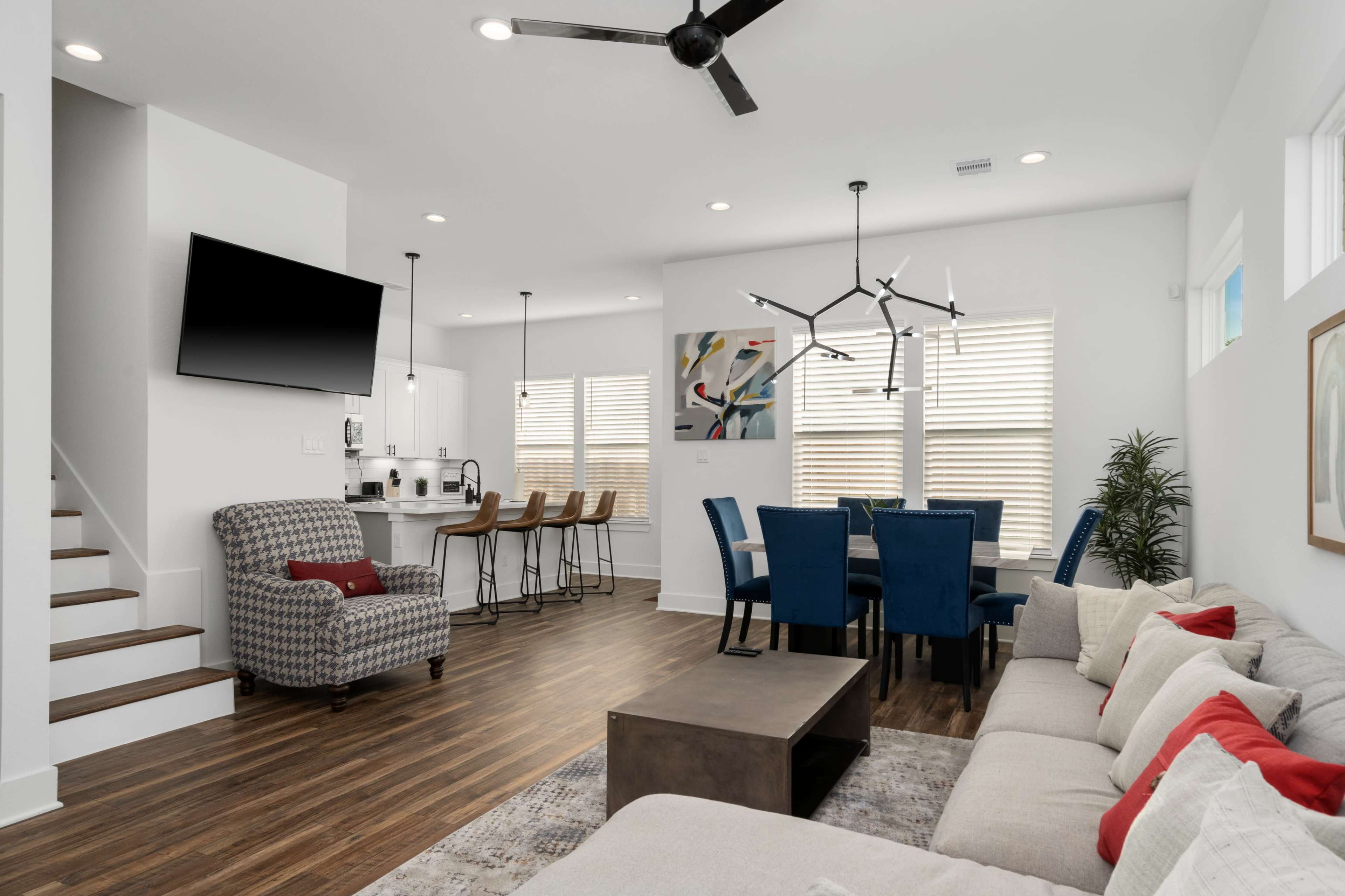 A modern living area with a sectional sofa, a coffee table, a dining space with blue chairs, and a kitchen bar with stools, all featuring ample natural light.
