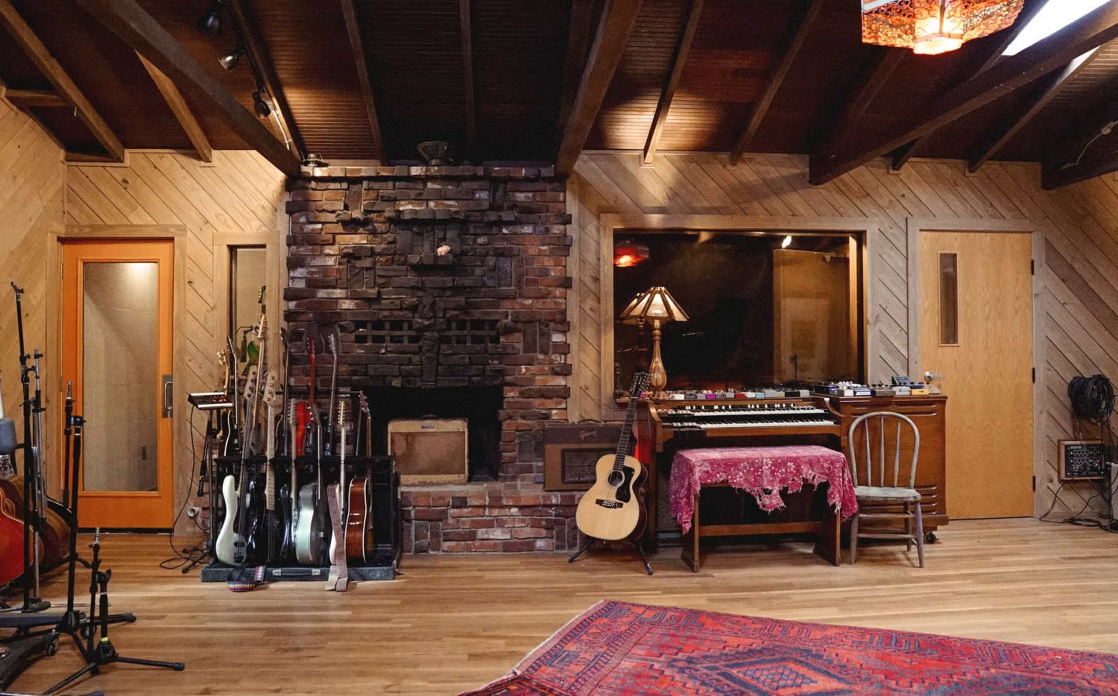 The image shows a rustic music studio featuring a stone fireplace, various guitars on stands, a vintage synthesizer on a wooden table, and wooden walls with large windows.