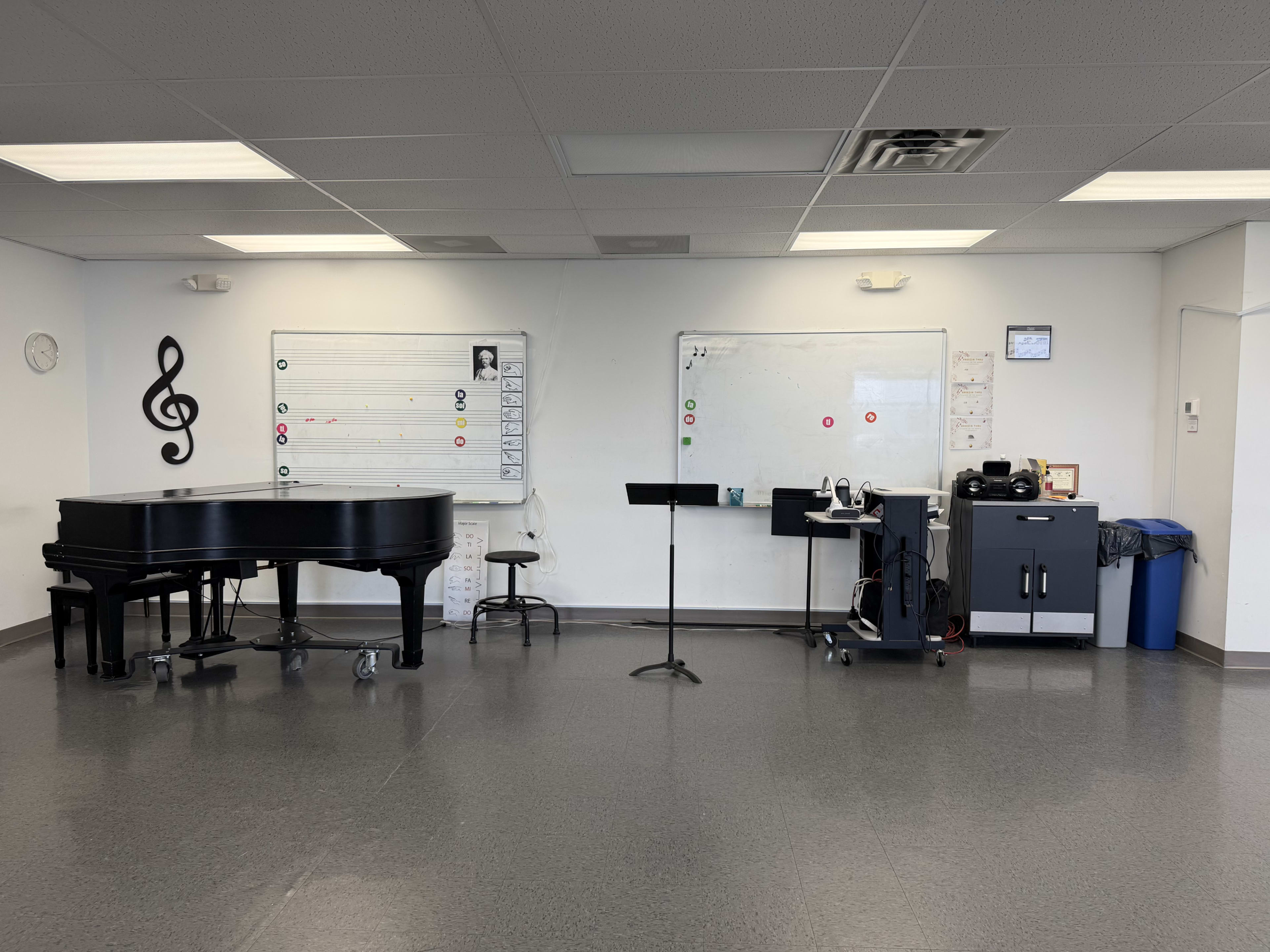 The image shows a music classroom featuring a grand piano, a music stand, a rolling cart with equipment, and whiteboards on the walls.