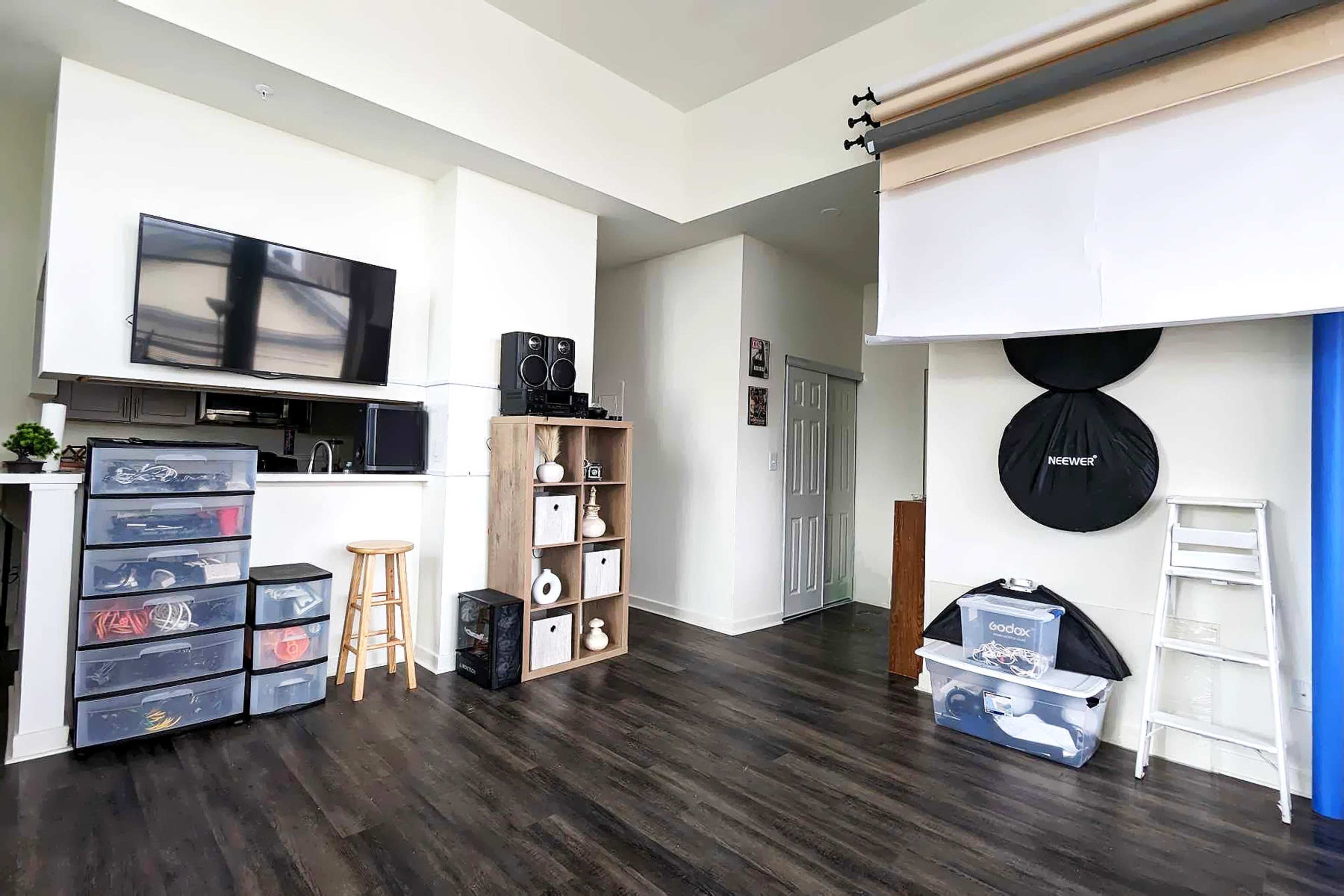 A bright room featuring a kitchenette with a television, storage bins, a ladder, and various photography backdrops along one wall.