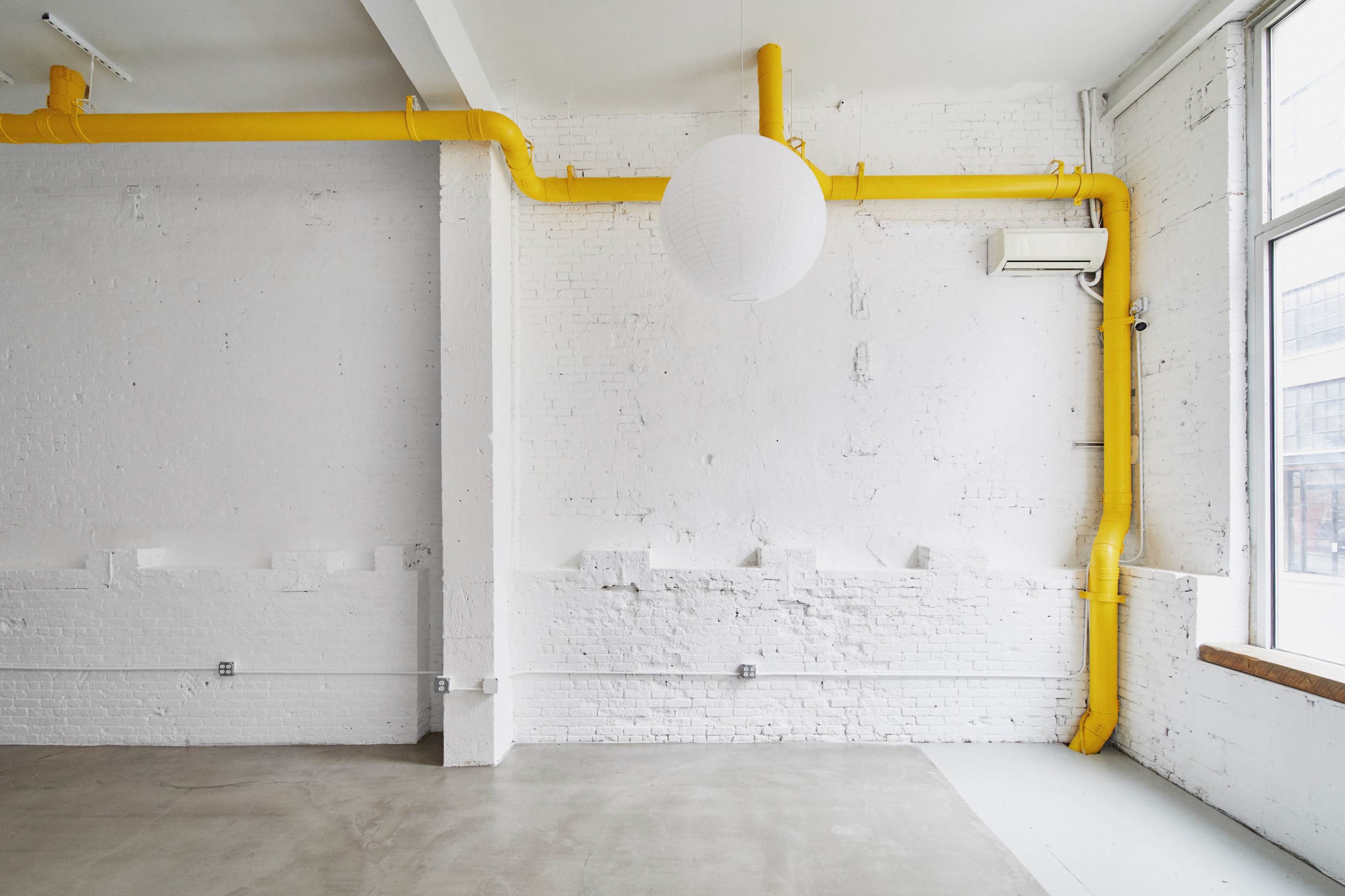A minimalist white room with exposed yellow piping along the wall and a round paper lantern hanging from the ceiling.