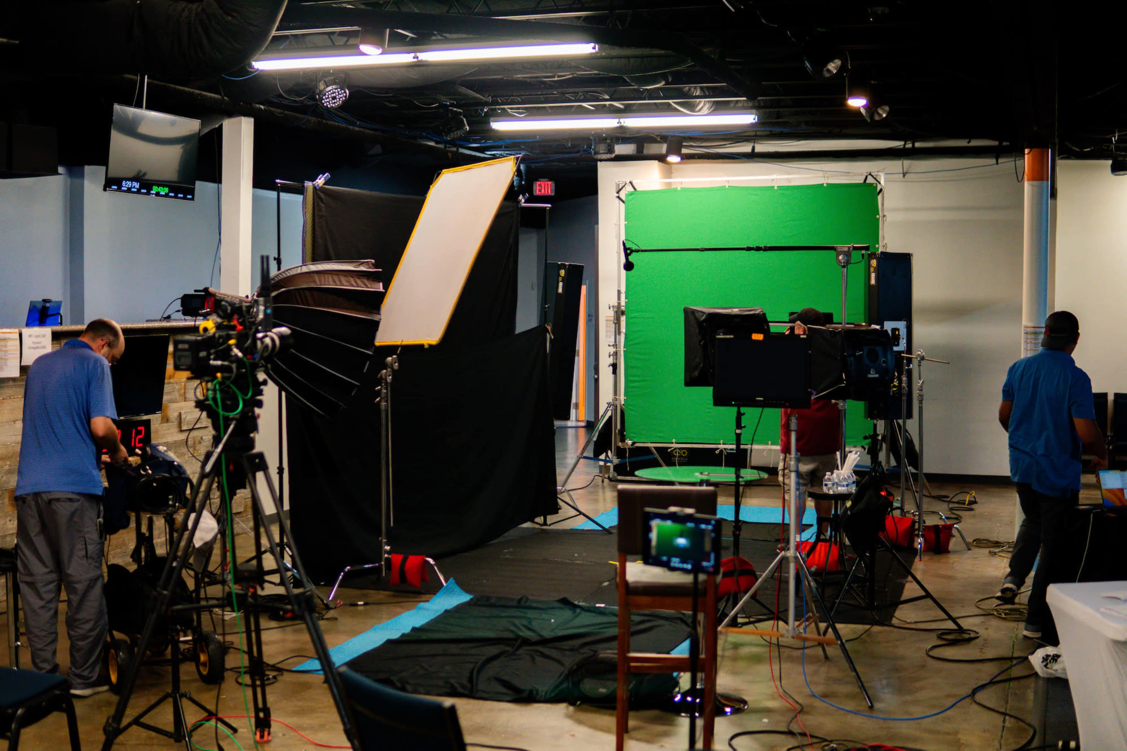 The image shows a film or photography studio setup with various lighting equipment, a green screen, and crew members preparing for a shoot.