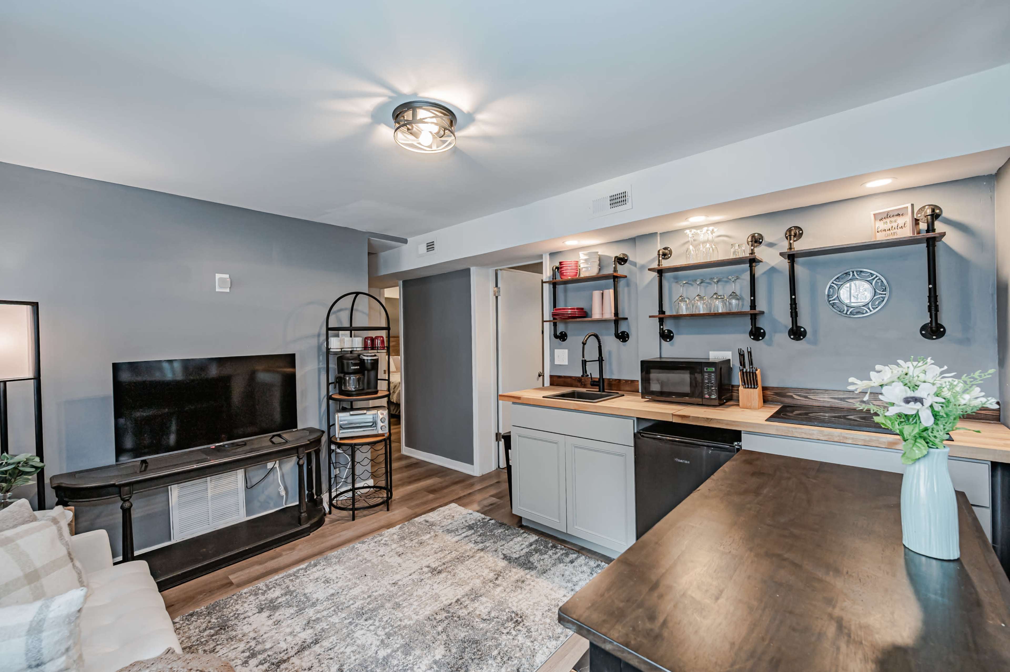 A modern, compact kitchen and living area featuring a television, a small countertop with a microwave, and shelves displaying glassware.