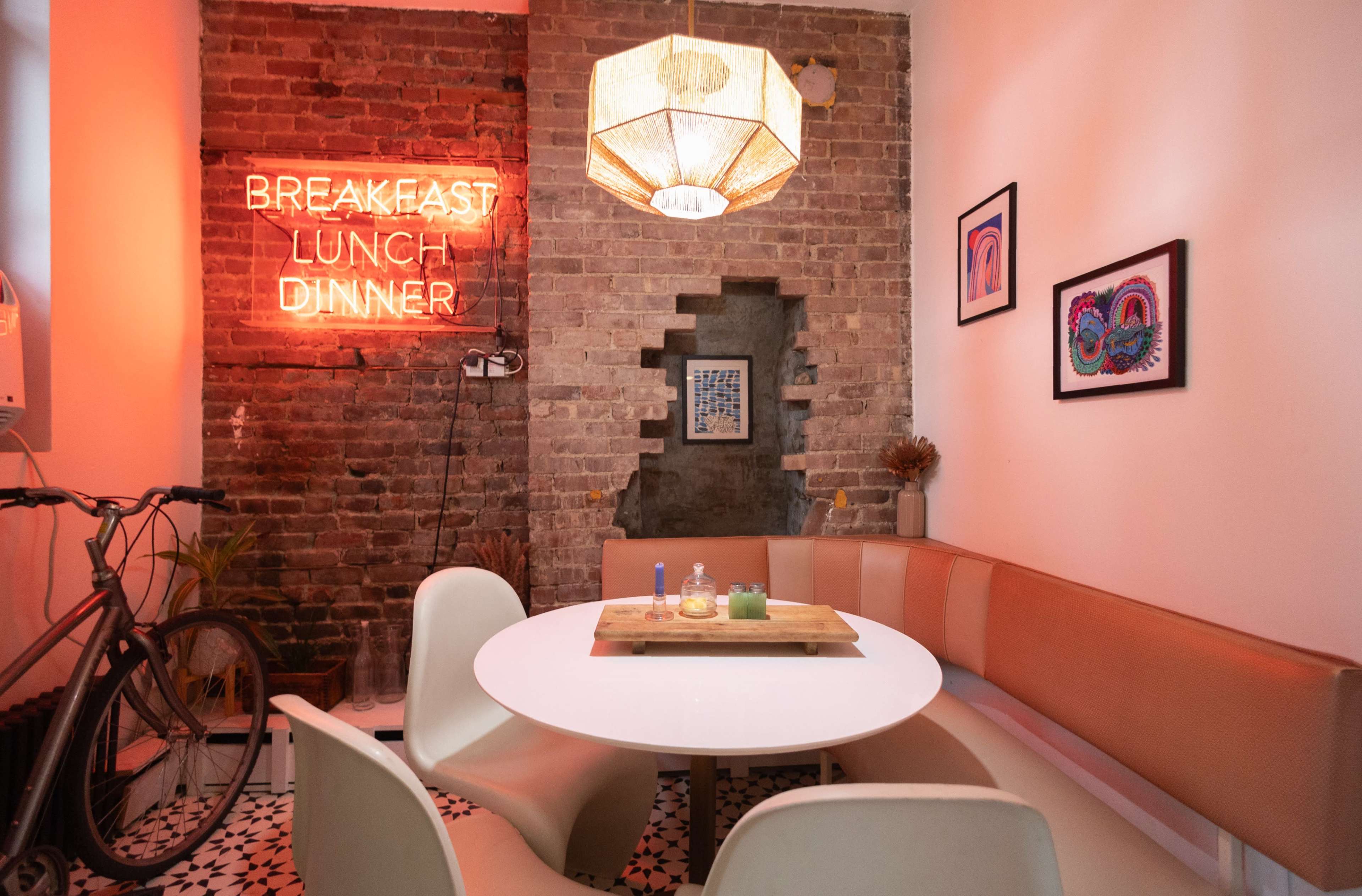 A cozy dining area features a round white table surrounded by curved orange benches, illuminated by a neon sign that reads "BREAKFAST LUNCH DINNER."