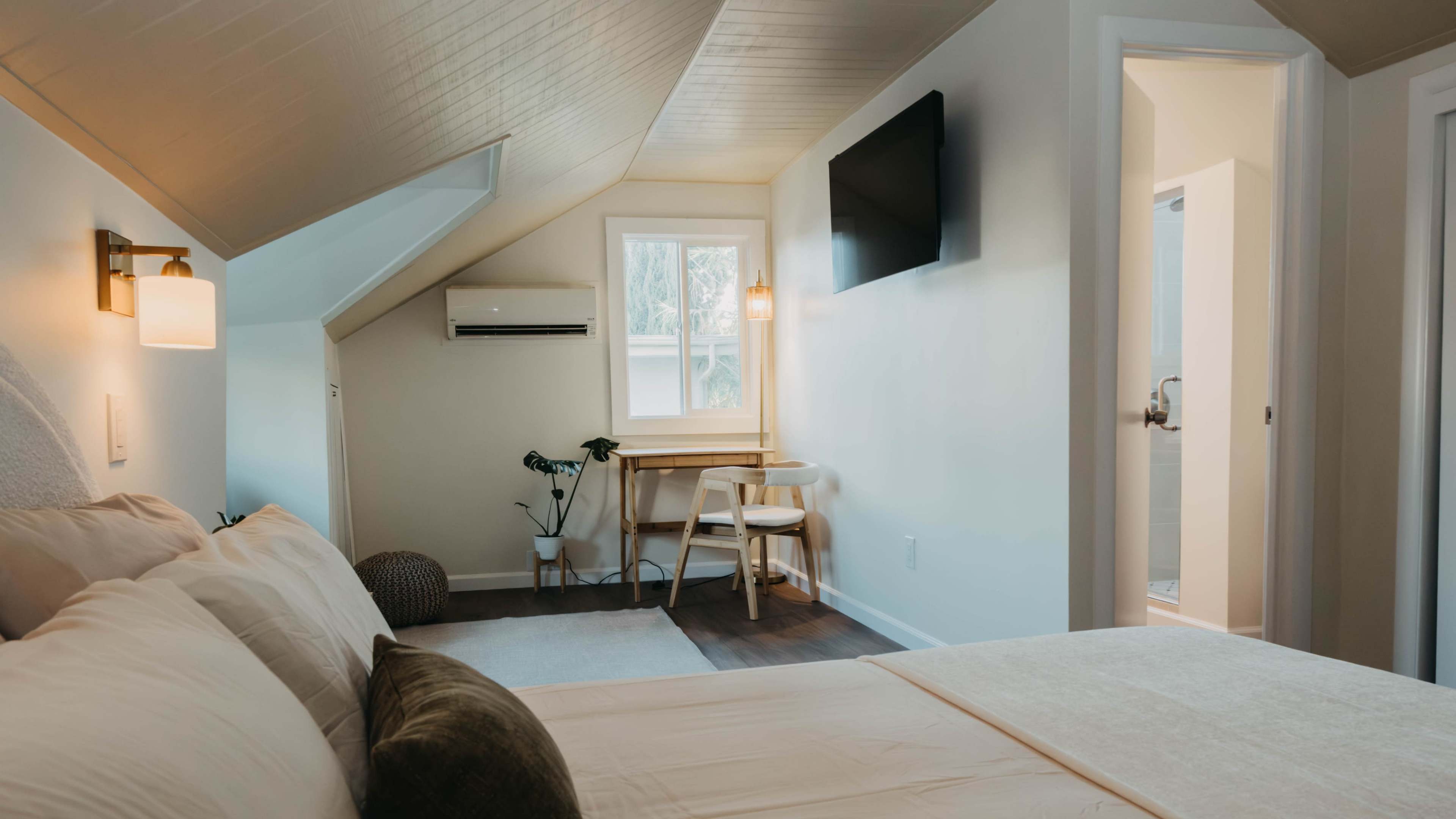The image shows a cozy bedroom with a sloped ceiling, a bed, a small table and chair, a window, and a wall-mounted television.